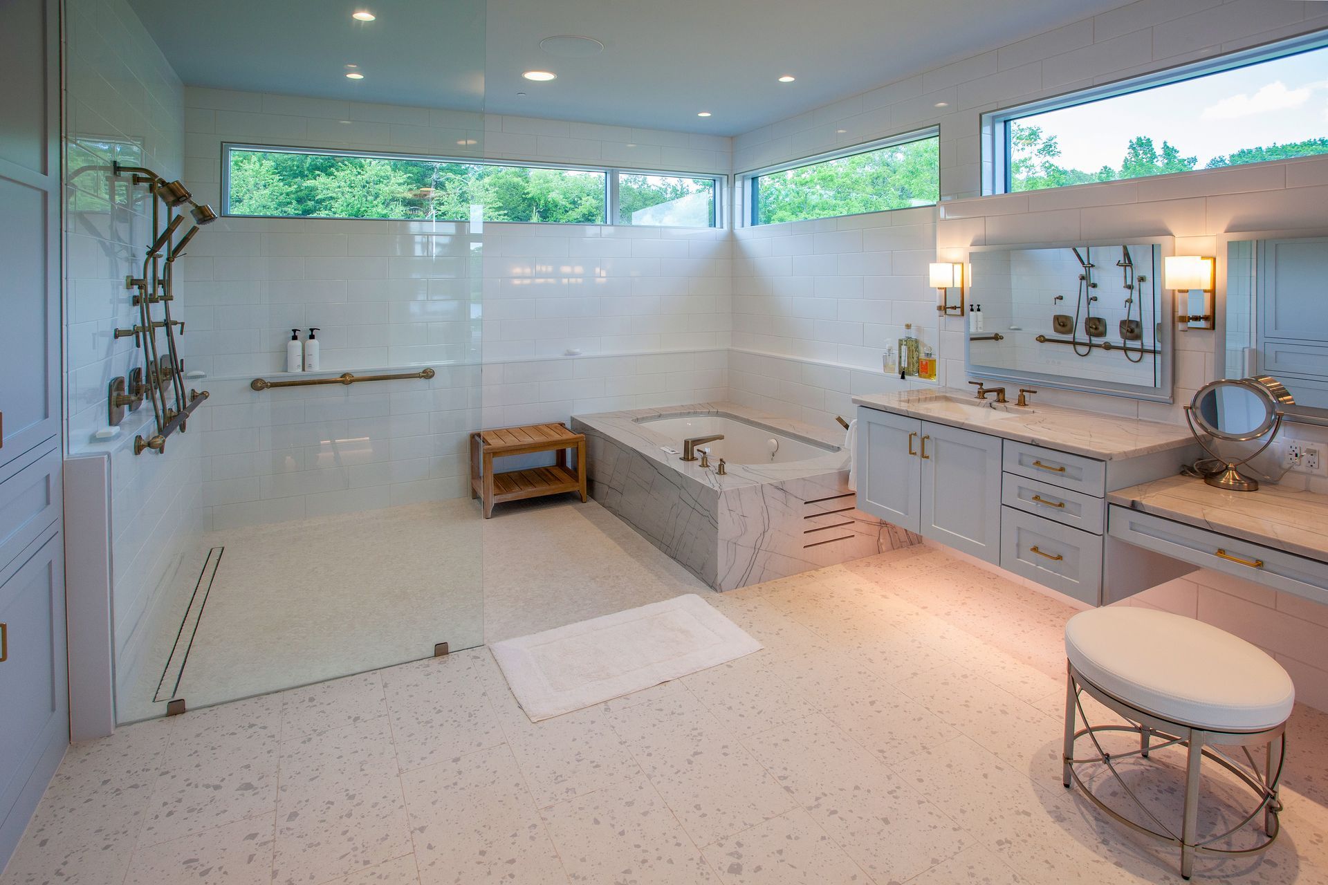 A bright, modern bathroom with a large walk-in shower, soaking tub, double vanity, and vanity stool on speckled flooring.