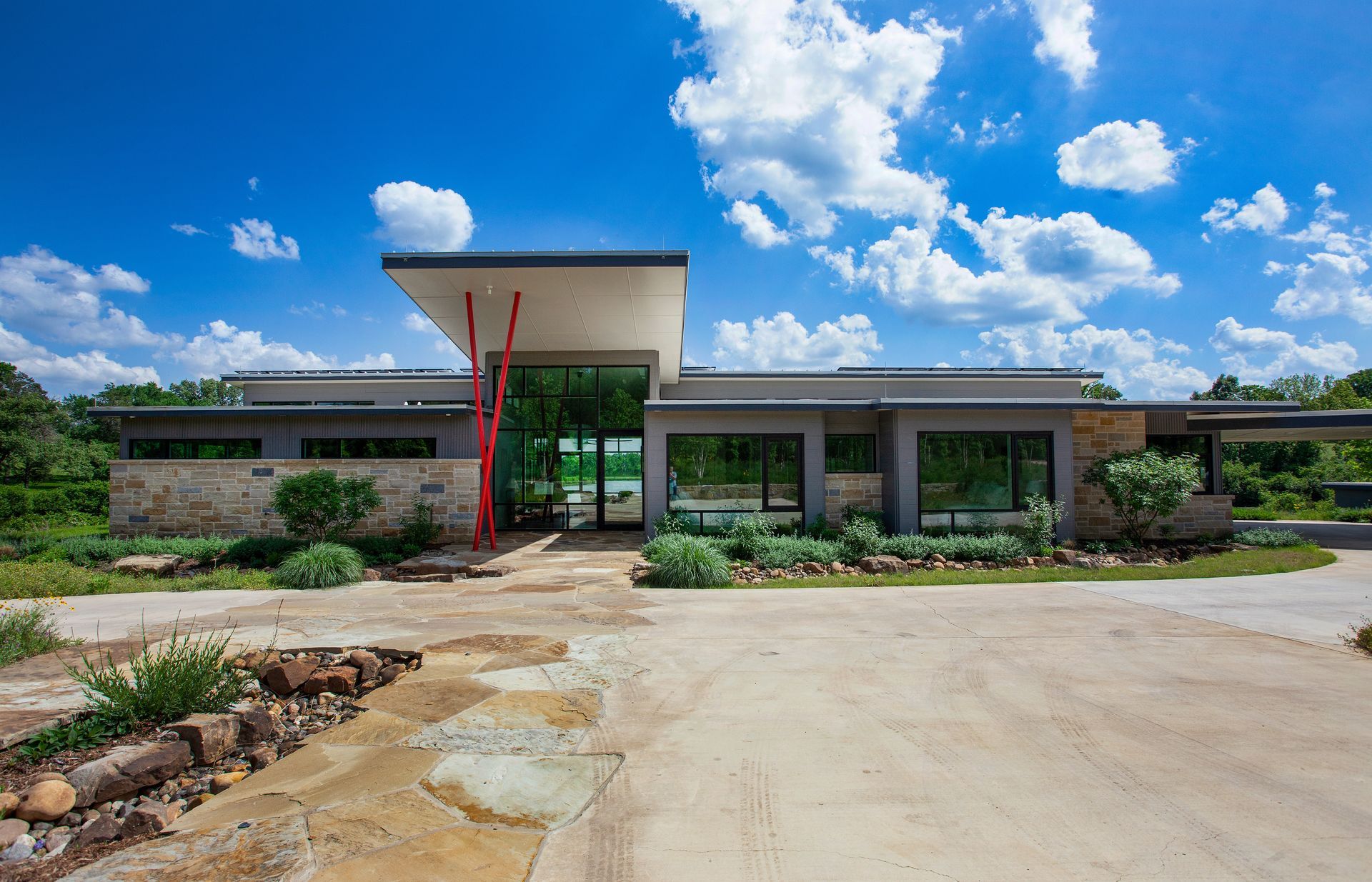 Modern single-story building with stone exterior, a flat roof, and a red support beam under a bright blue sky.