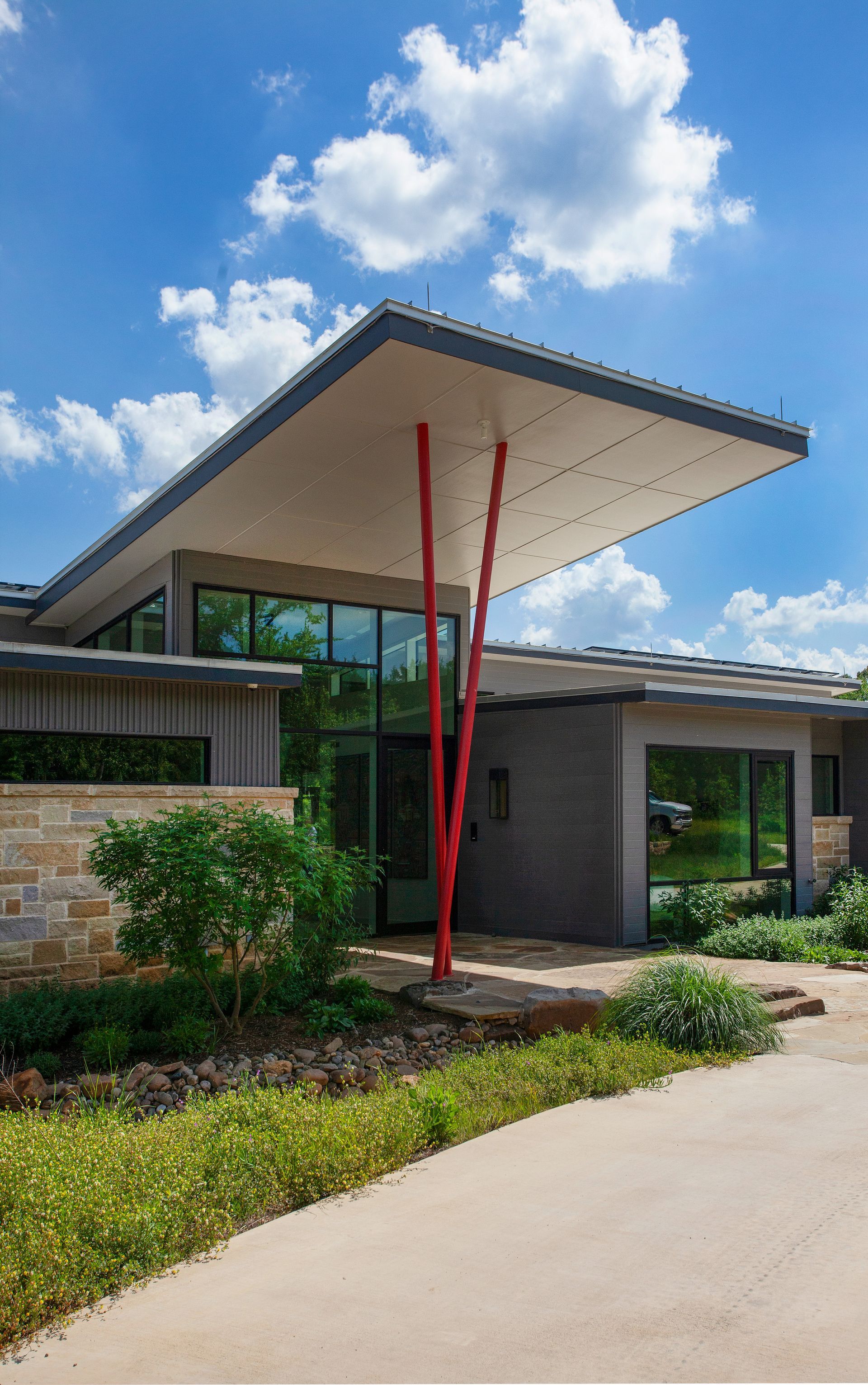 Modern home entrance with a large overhanging flat roof supported by two angled red metal pillars against a blue sky.
