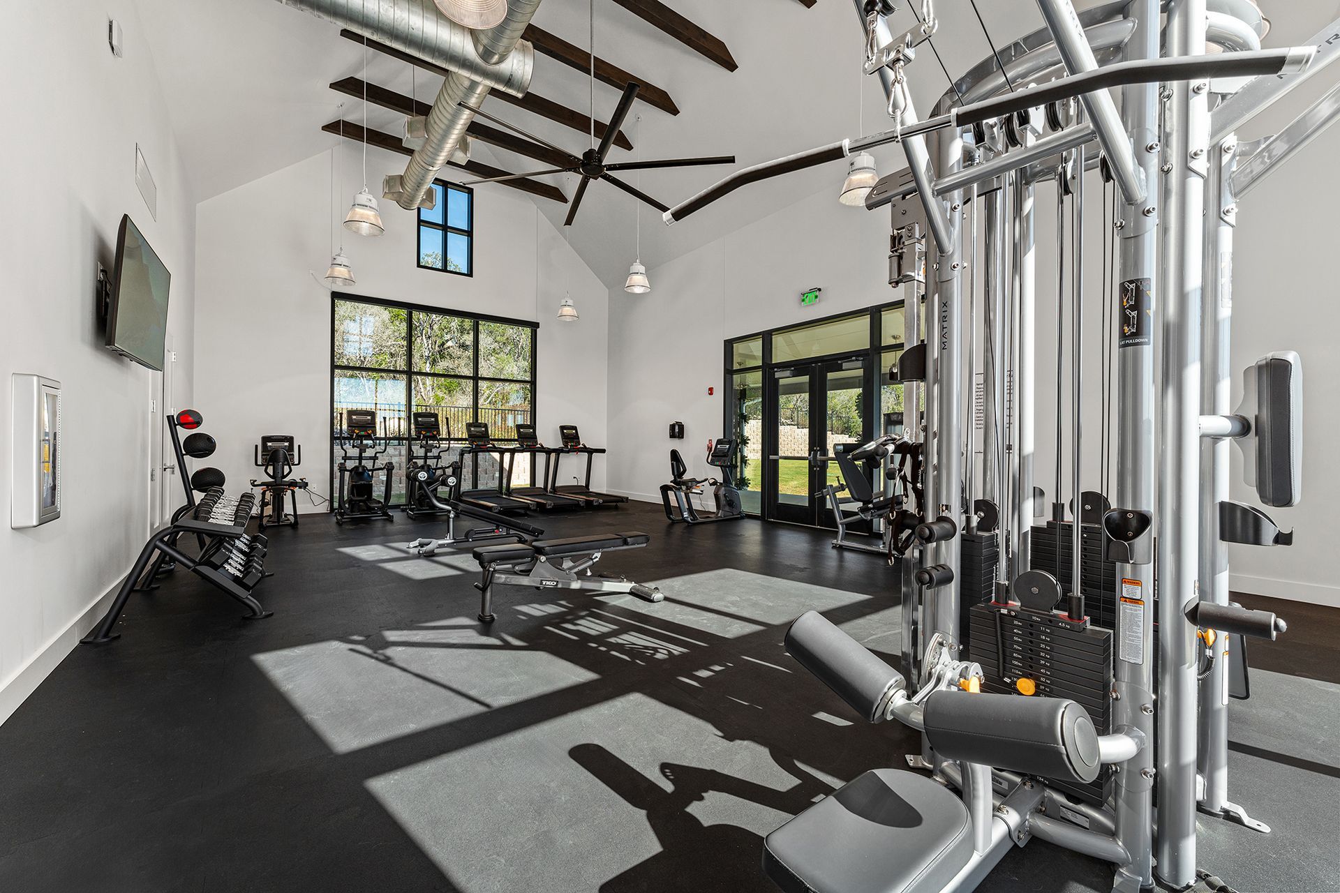 A bright, high-ceilinged fitness center with various workout machines, rubber flooring, and large windows.
