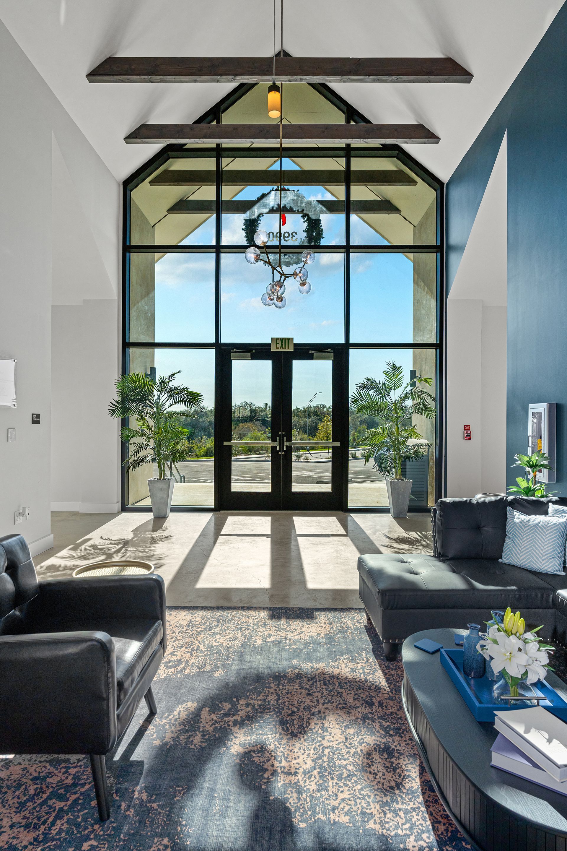 A bright, modern lobby with a high-ceiling glass entrance, dark furniture, and a decorative wreath hanging on the doors.