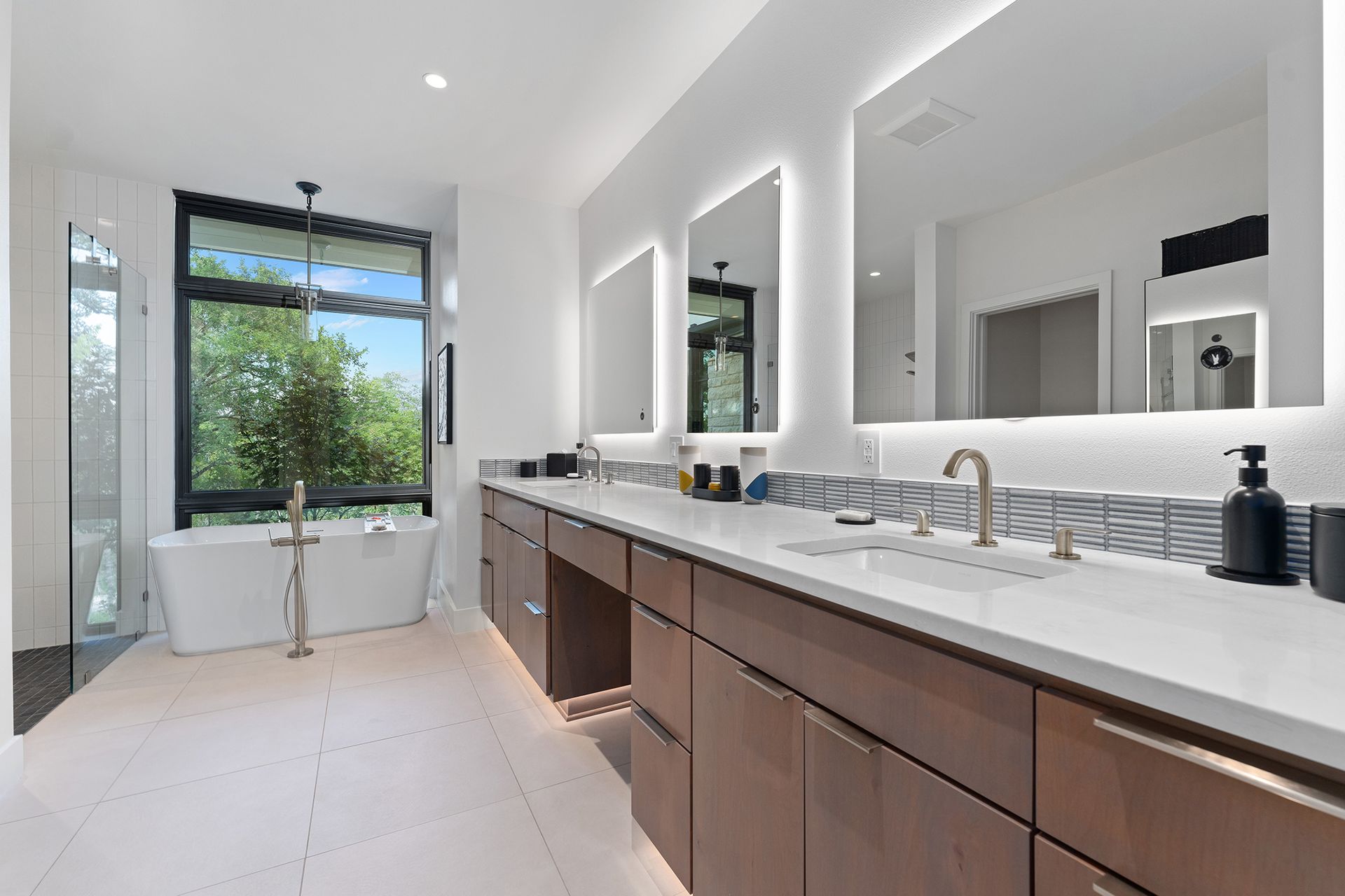 Modern bathroom featuring a long wood vanity, white countertop, two lighted mirrors, and a freestanding soaking tub.
