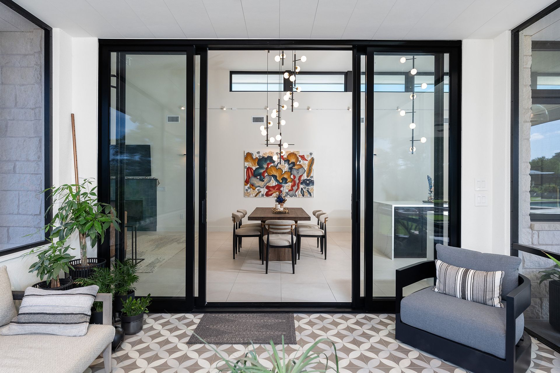 A patio overlooking a bright modern dining area with a chandelier, wooden table, and art, framed by black glass doors.