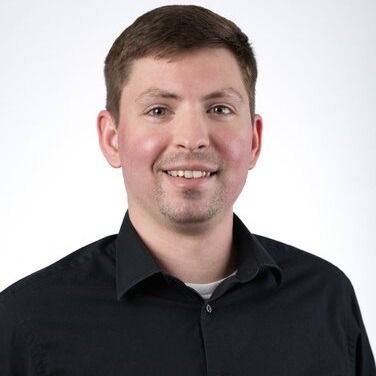 A person with light brown hair and a short beard wearing a black button-down shirt, smiling against a white background.