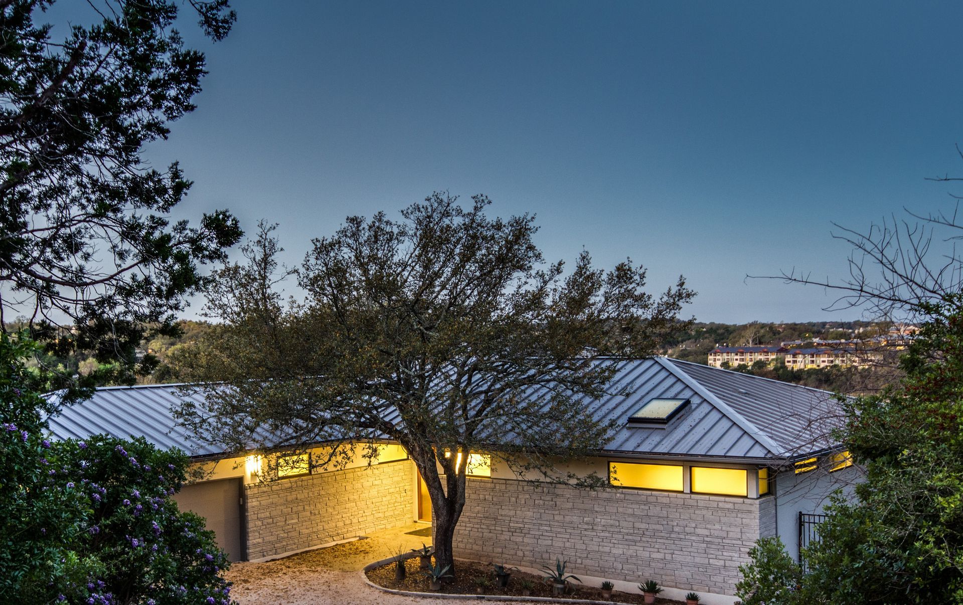 A modern stone house with a metal roof and glowing warm interior lights, nestled among trees during twilight.