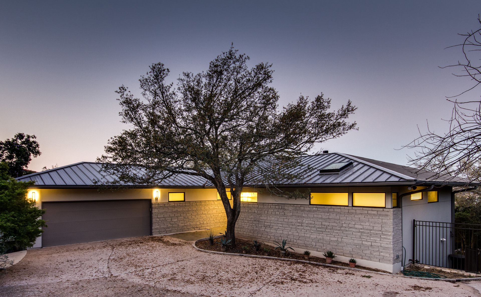 A modern, single-story house with a metal roof and light stone facade, set behind a large tree at dusk.