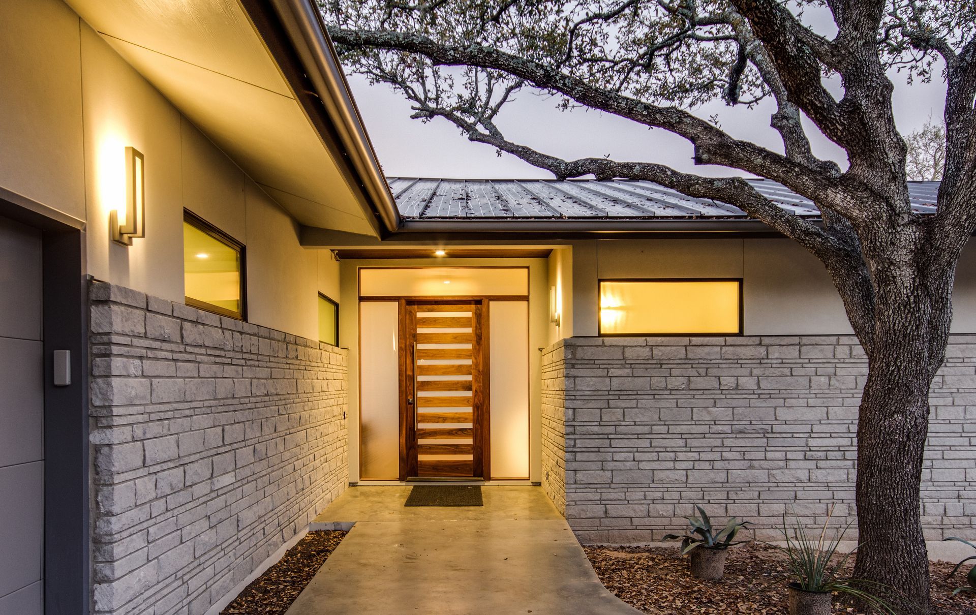 Modern home entrance with a wood-slatted door, horizontal stone walls, and warm exterior lighting at dusk.