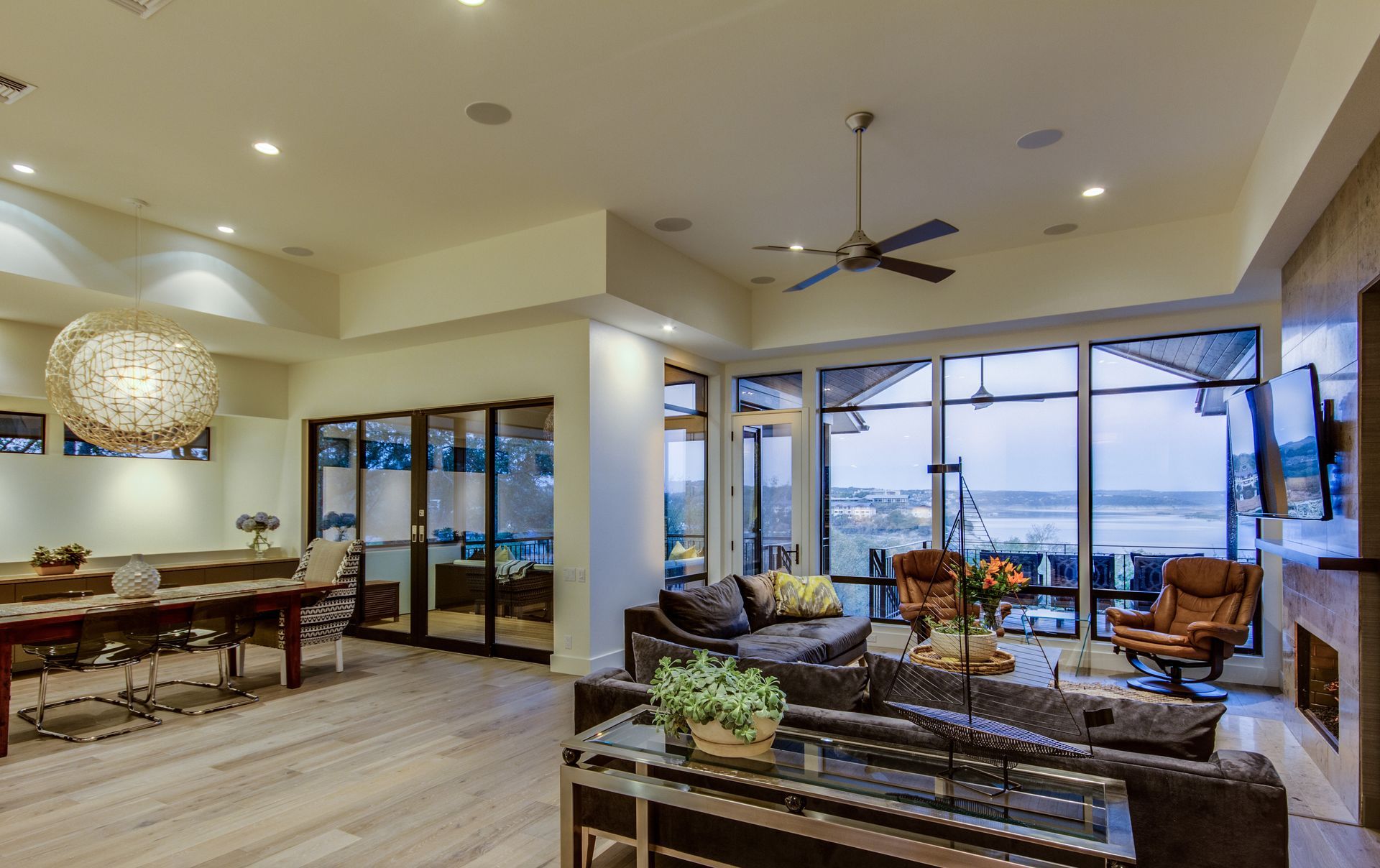 Modern, open-concept living area with a large dining table, gray seating, and expansive windows overlooking a water view.