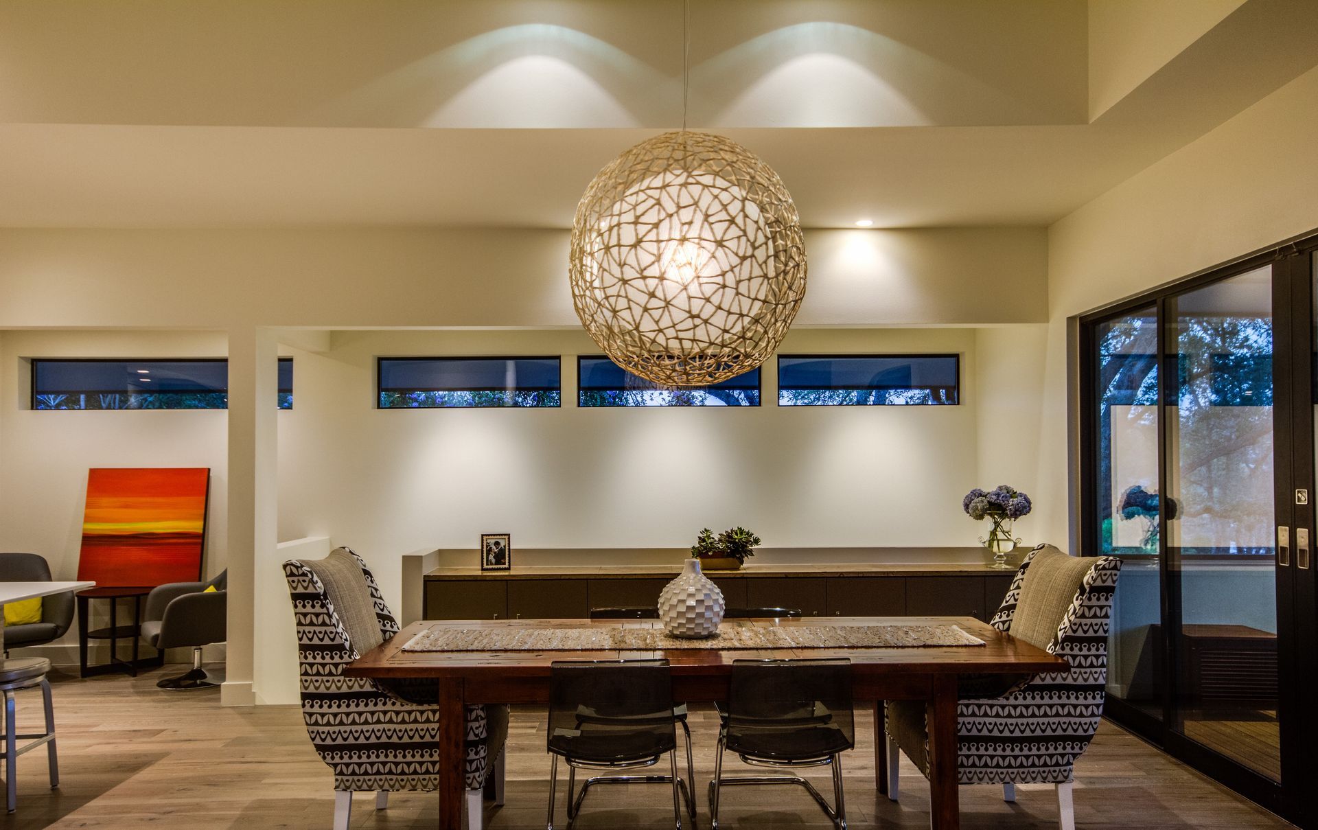 A dining room with a large, spherical woven pendant light above a wooden table, patterned chairs, and horizontal windows.
