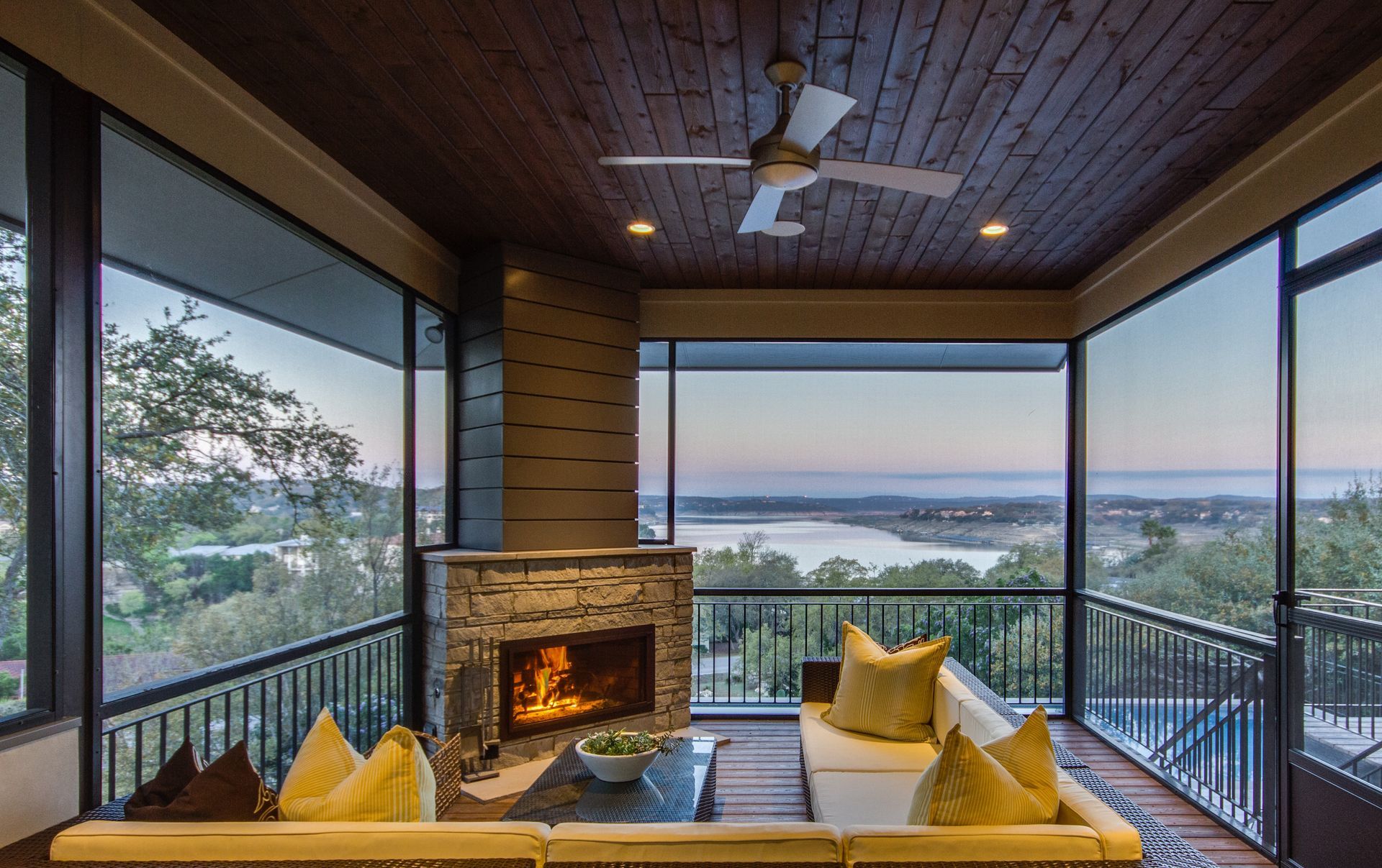 A cozy screened-in porch with a fireplace, comfortable seating, and a scenic view of a lake at dusk.