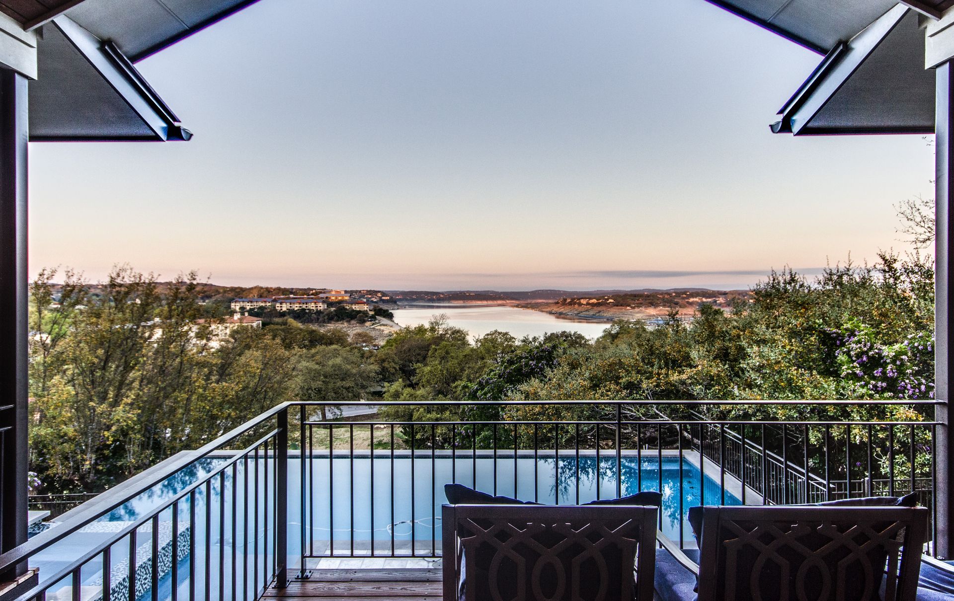 A balcony overlooking a swimming pool, trees, and a river under a clear sky.