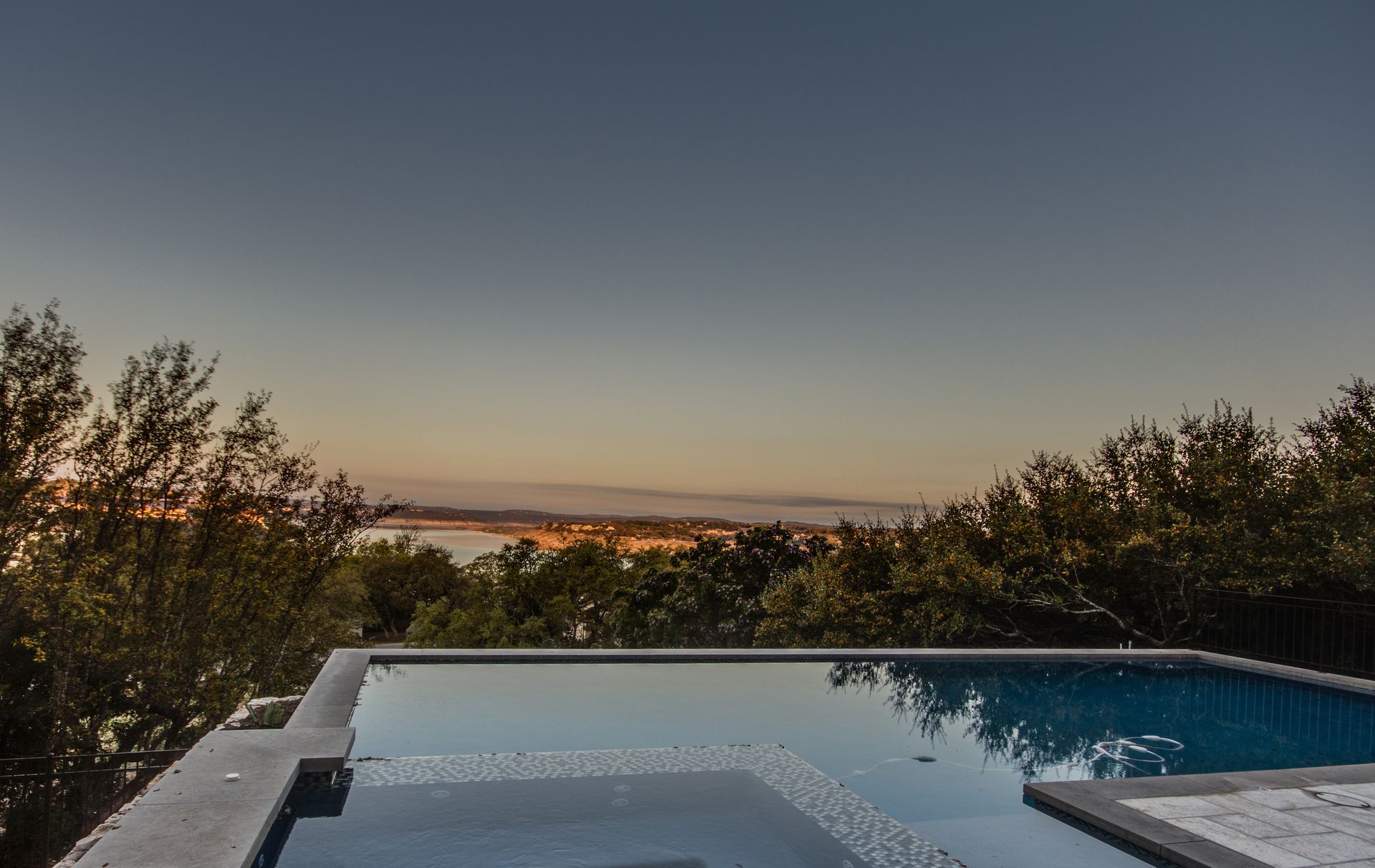 Infinity pool overlooking a sunset view of a coastal landscape with trees in the foreground.