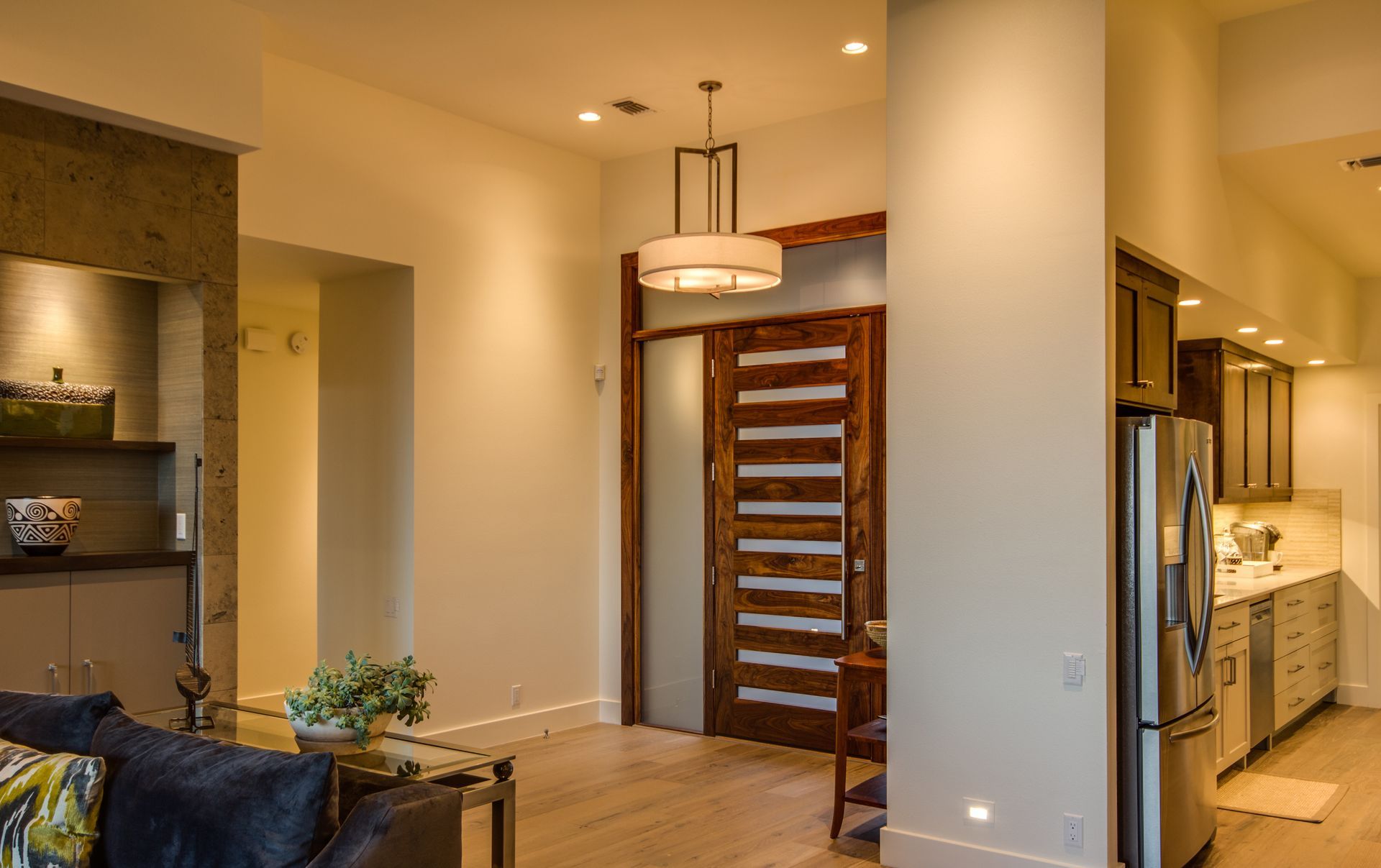 Modern living room featuring a wood-slat front door, a hanging light fixture, and an open kitchen with stainless appliances.