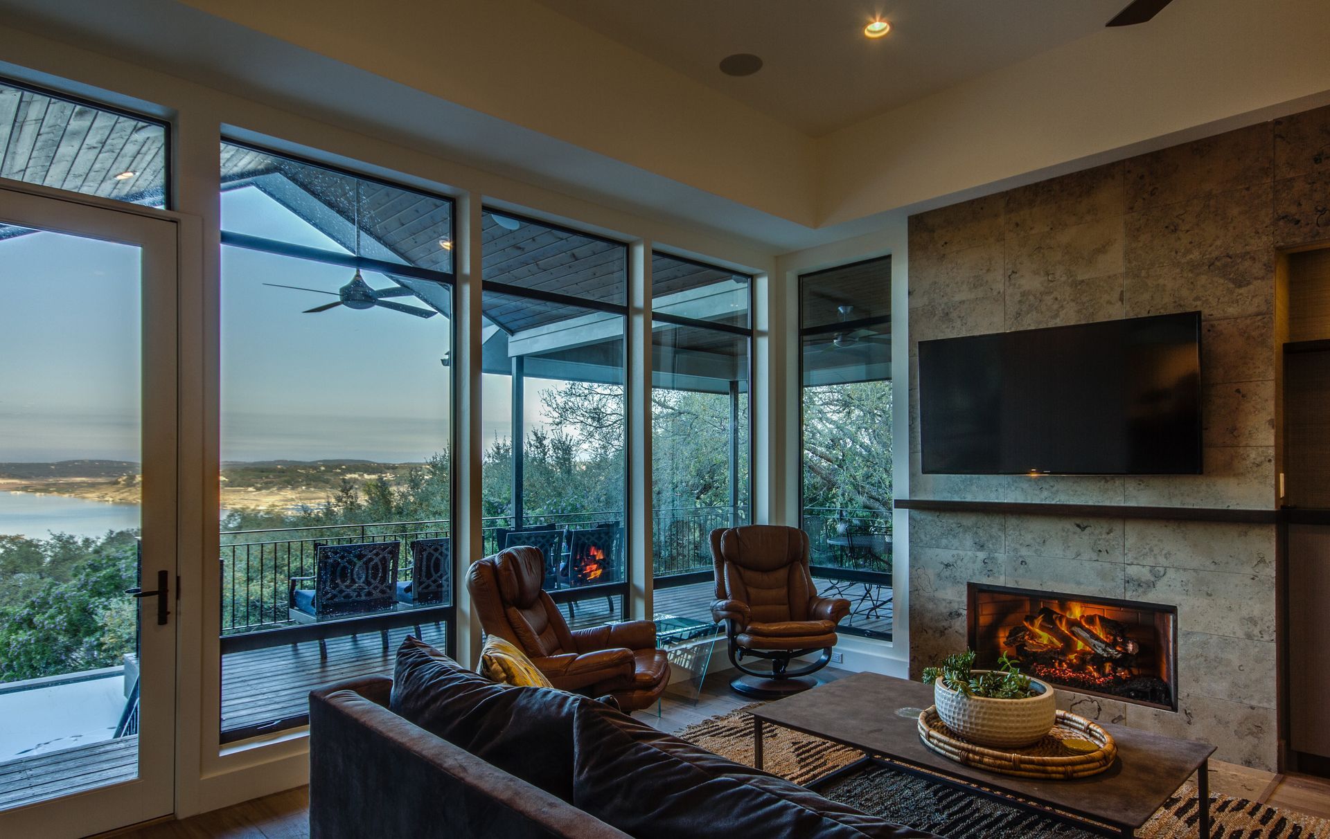 A cozy living room with a stone fireplace, flat-screen TV, armchairs, and floor-to-ceiling windows overlooking a lake.