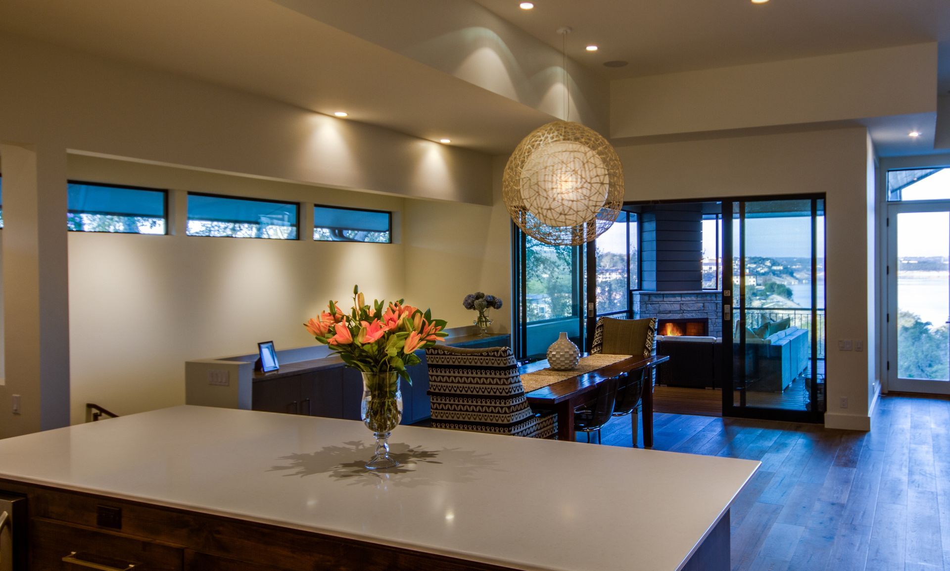 A kitchen island with a flower vase overlooks a modern dining area and a patio with a fireplace at dusk.