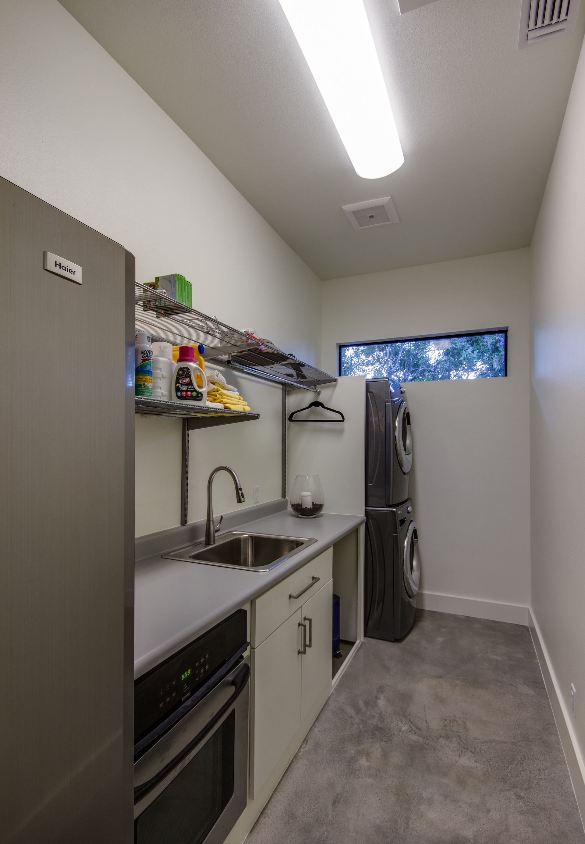 A laundry room with a grey countertop, sink, lower cabinets, open shelving, and a stacked washer and dryer unit.