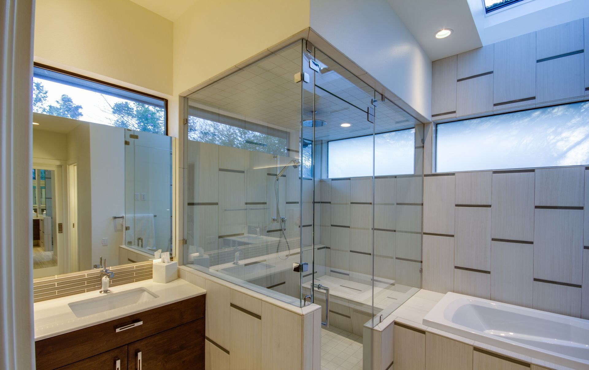 A modern bathroom with a wood vanity, a glass-enclosed shower, and a white soaking tub set against tile walls.