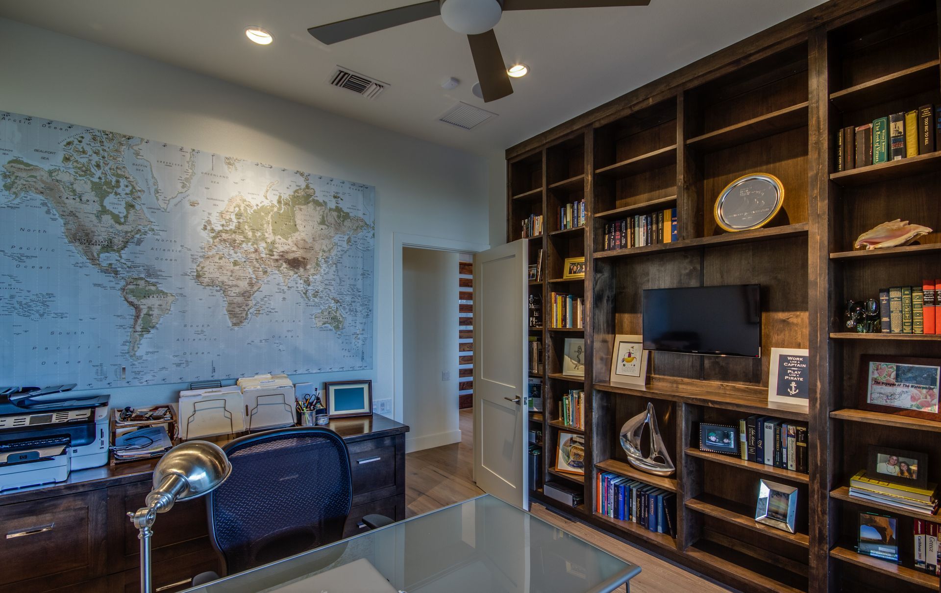 Home office with a large world map mural, a dark wood bookcase wall, and a desk with a computer and lamp.