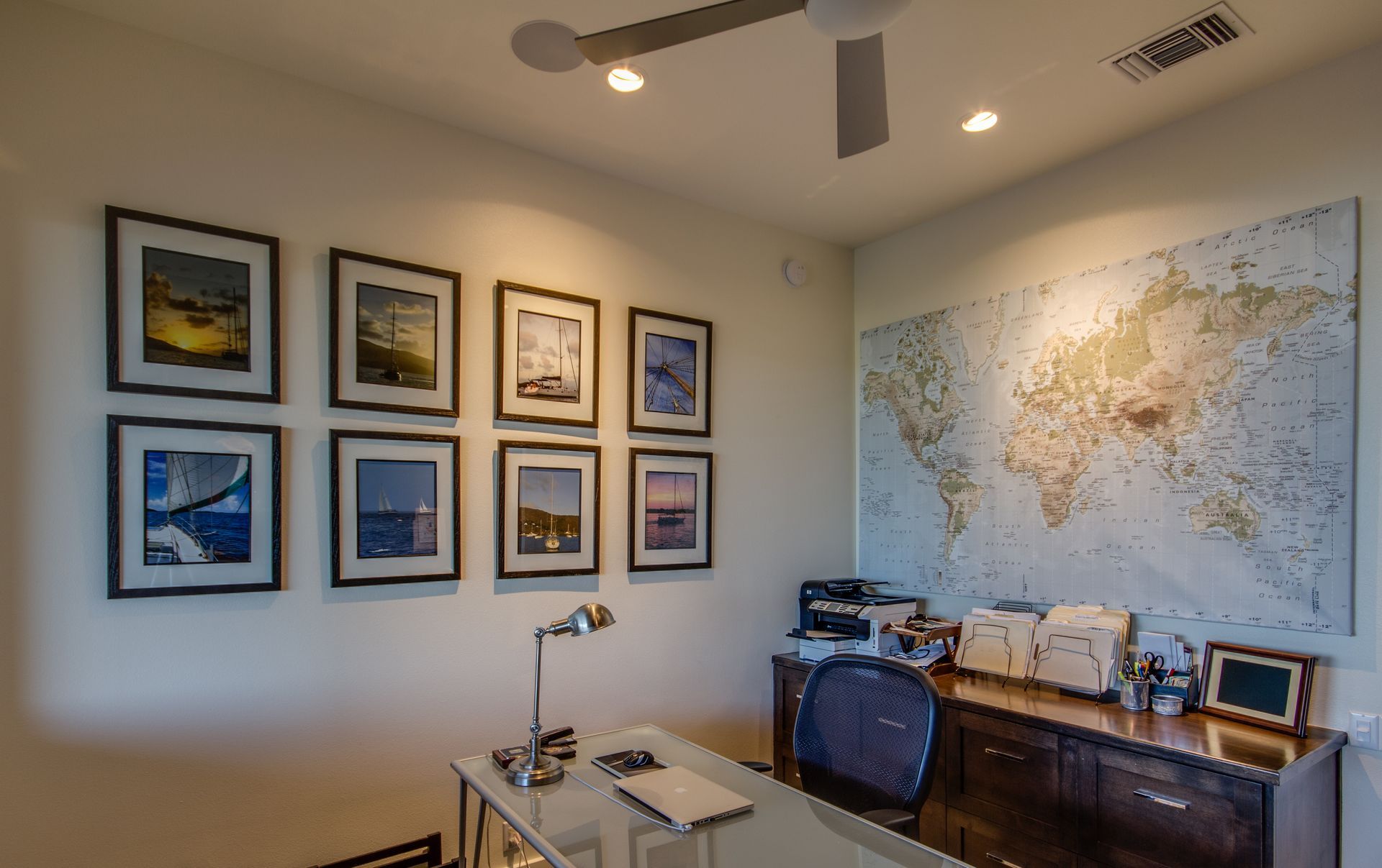 A home office with a glass desk, a dark chair, a large world map on the wall, and eight framed pictures arranged in grids.