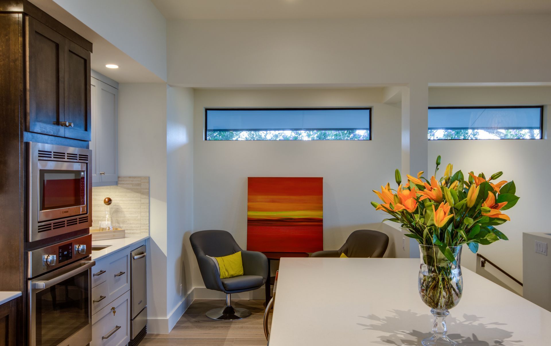 A kitchen with a dark-wood oven cabinet, a white island, orange artwork, and two chairs under horizontal windows.