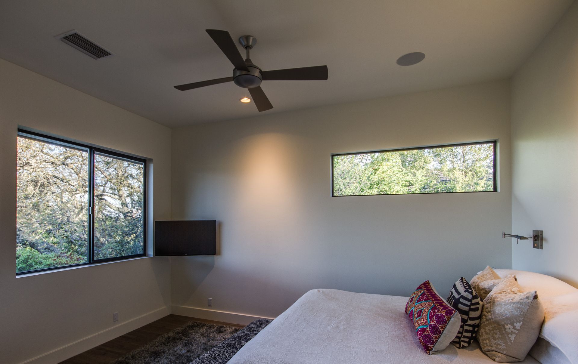 A modern bedroom with a bed featuring patterned pillows, a ceiling fan, a wall-mounted TV, and windows looking outside.