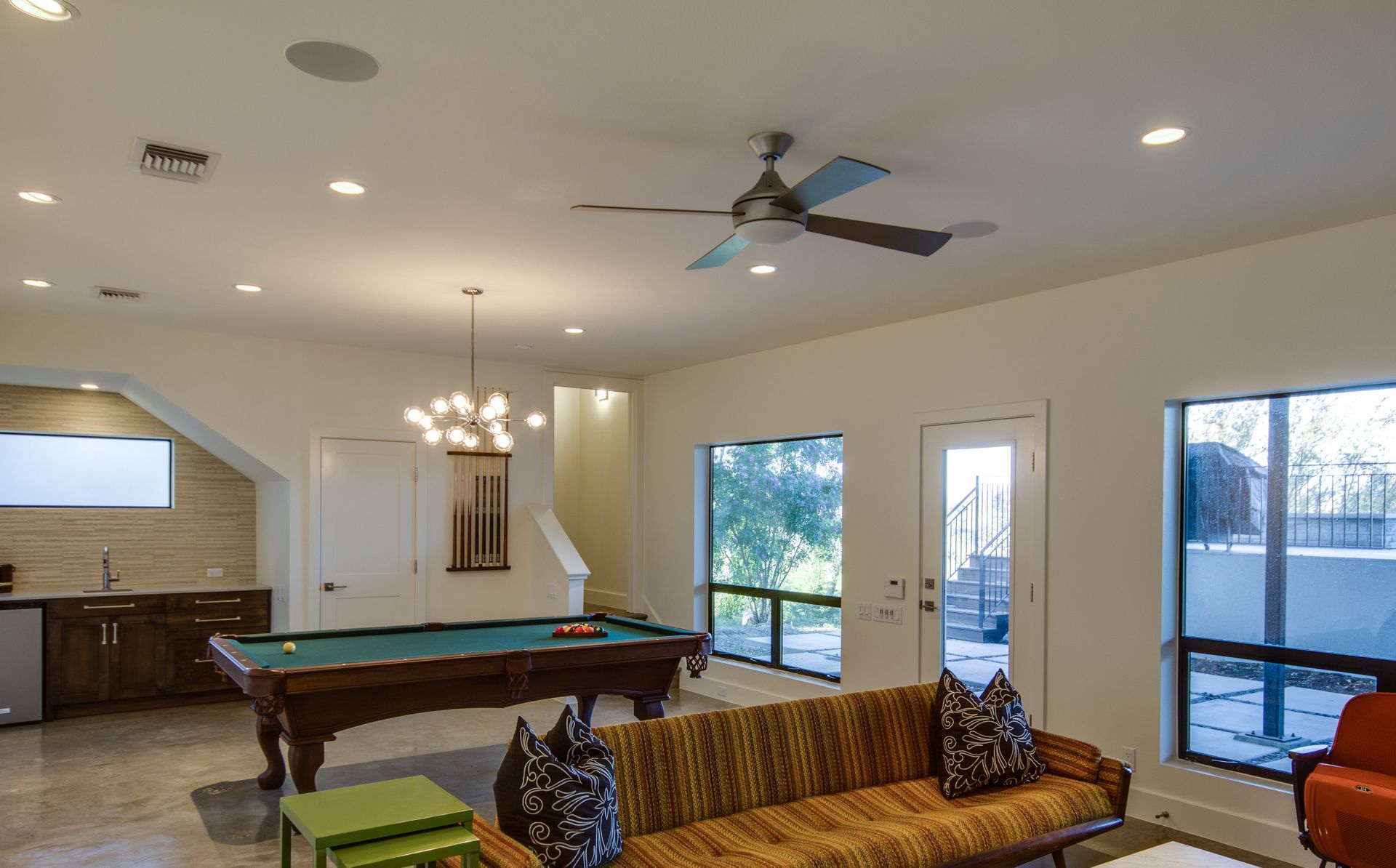 A spacious game room with a pool table, a patterned sofa, a modern chandelier, and large windows.