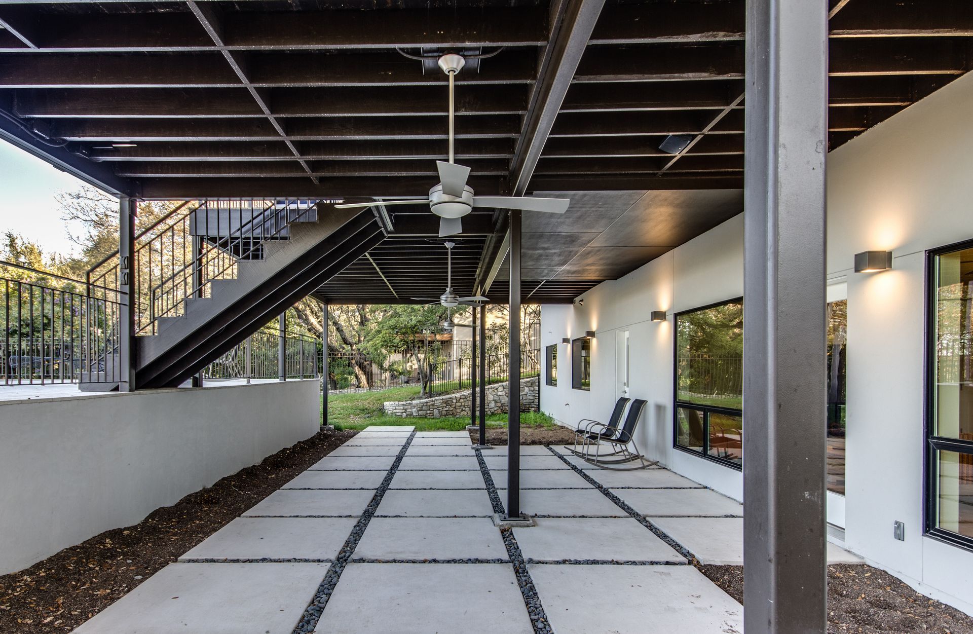 A covered concrete patio with a grid of pavers, stairs leading to an upper level, a ceiling fan, and modern wall lighting.