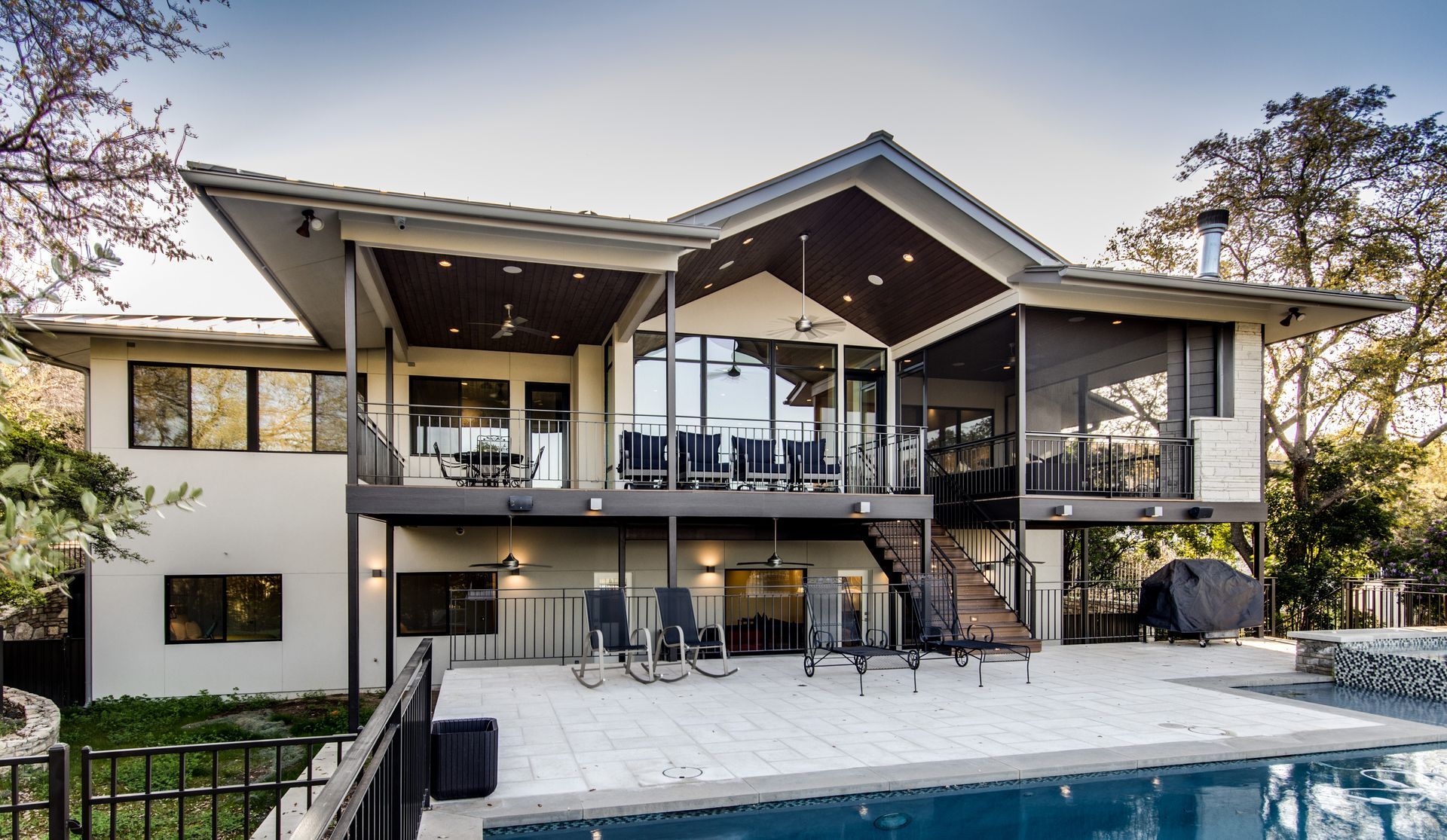 A two-story contemporary home with a large elevated deck, patio area, and a swimming pool in a wooded backyard.