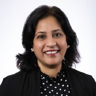 A smiling person with dark hair, wearing a black blazer over a black-and-white polka-dot top against a white background.