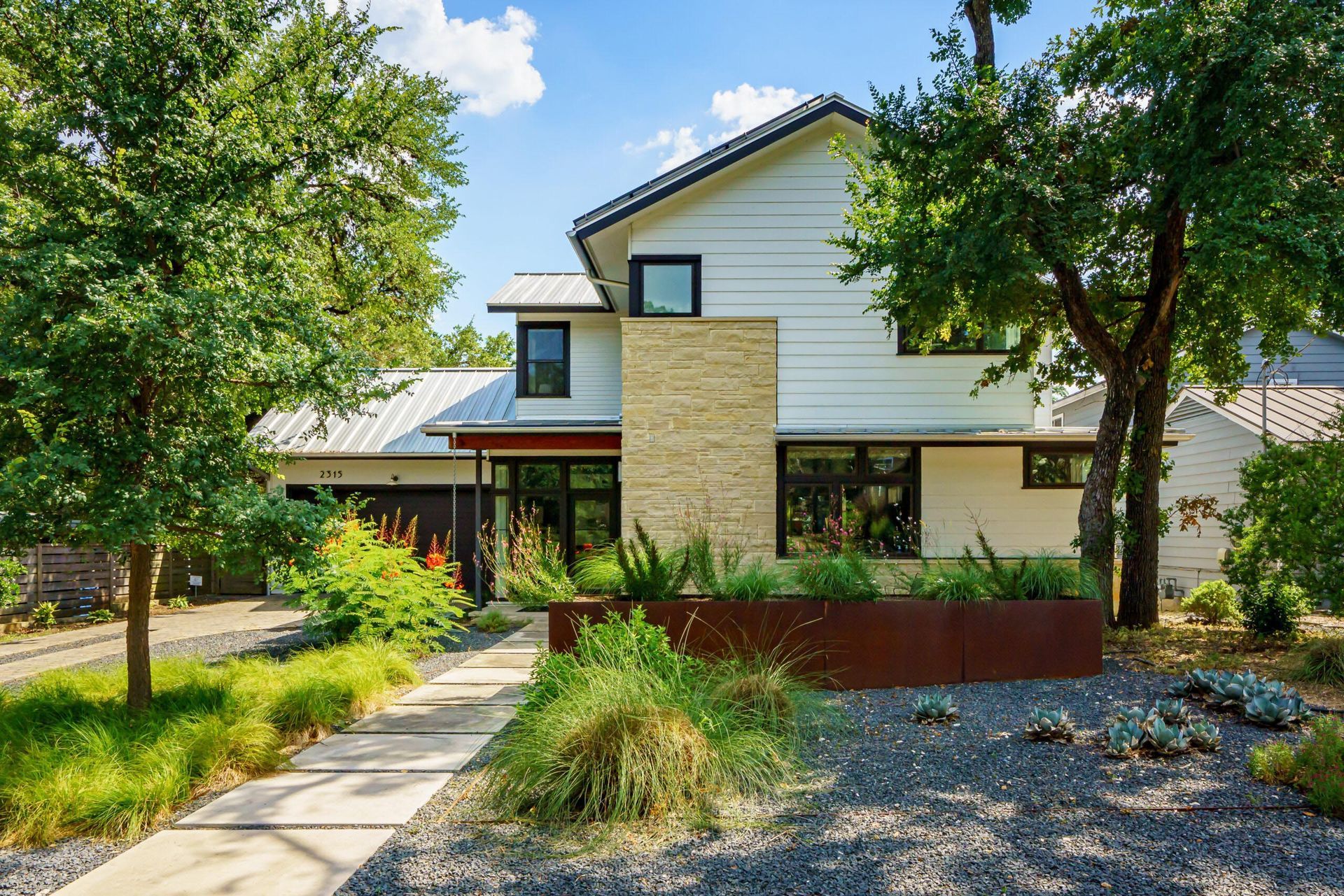 Modern white house with a stone accent and metal roof, with lush landscaping.
