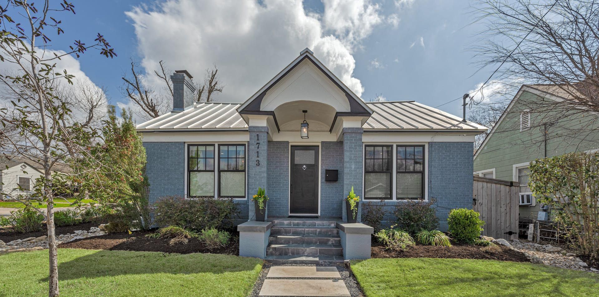 Front view of a small blue house with a white roof, centered walkway, and trimmed bushes