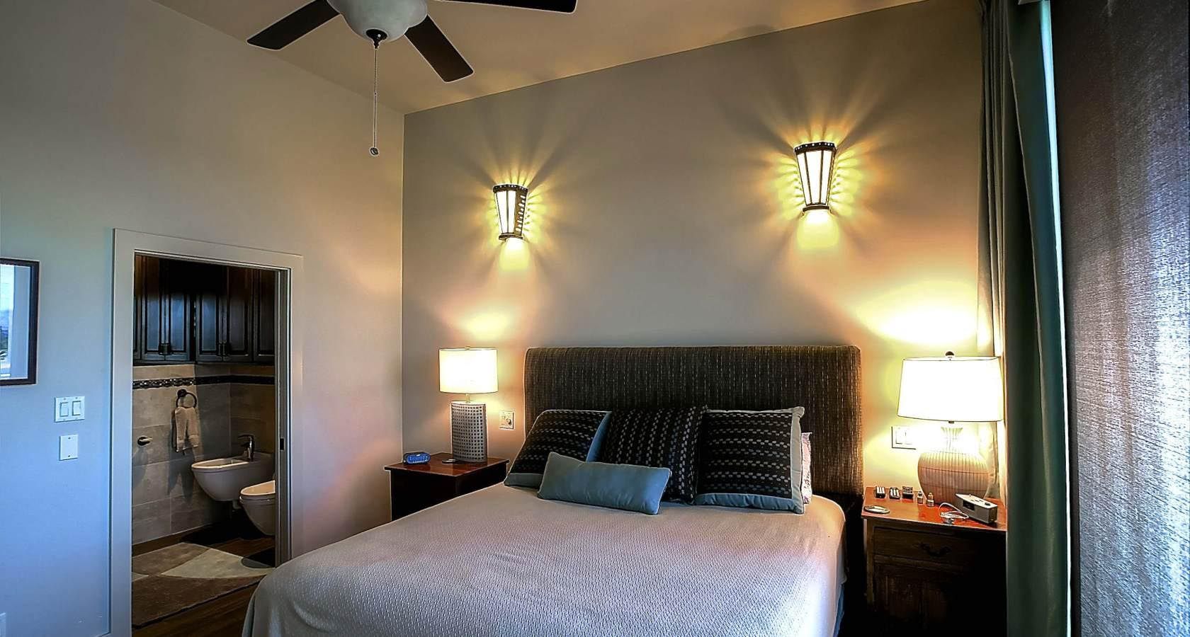 Bedroom with a bed, lamps, and sconces. Door to bathroom visible. Light beige walls.