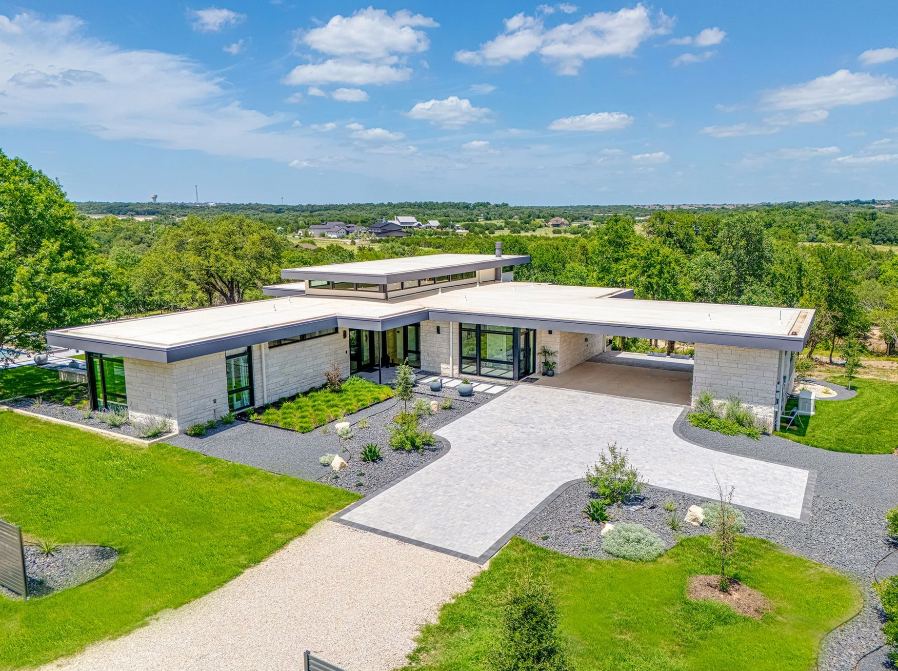 A modern, flat-roofed stone house with a gravel driveway set in a lush green landscape under a bright, cloudy sky.