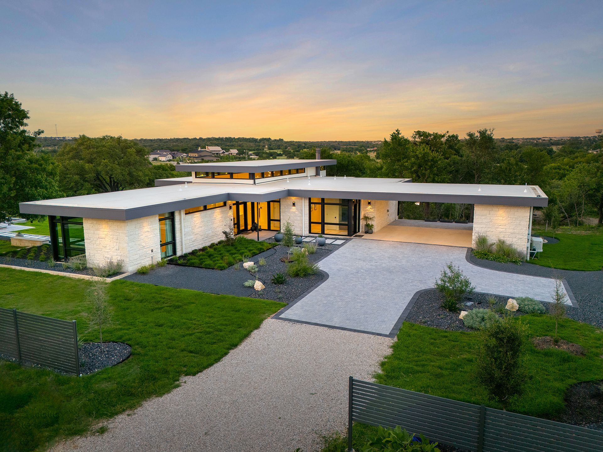 Modern home with a flat roof, white stone exterior, and gravel driveway at sunset.