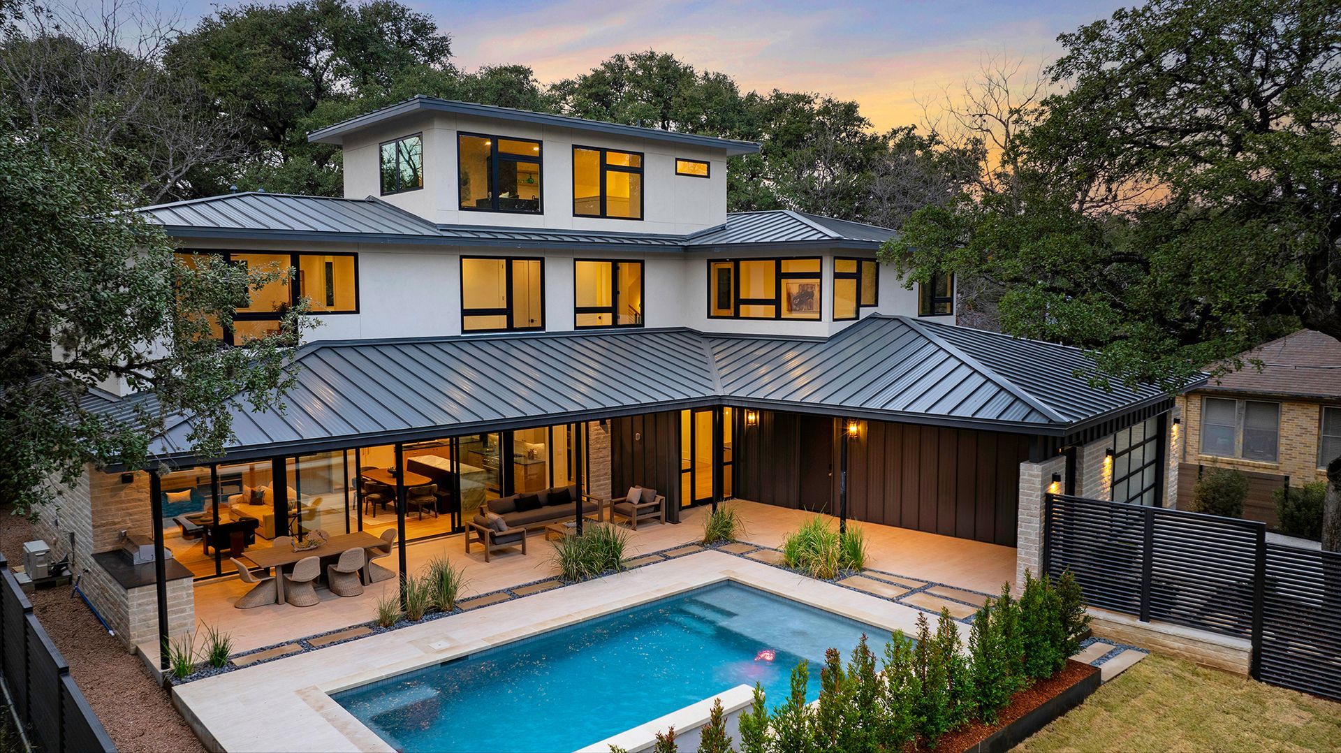 A modern white house with a dark metal roof, large windows, and a patio area overlooking a turquoise swimming pool.