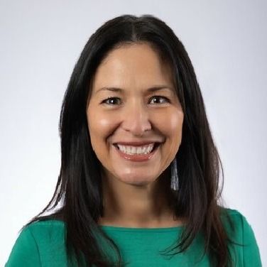 A smiling person with shoulder-length dark hair wearing a green top against a plain light background.