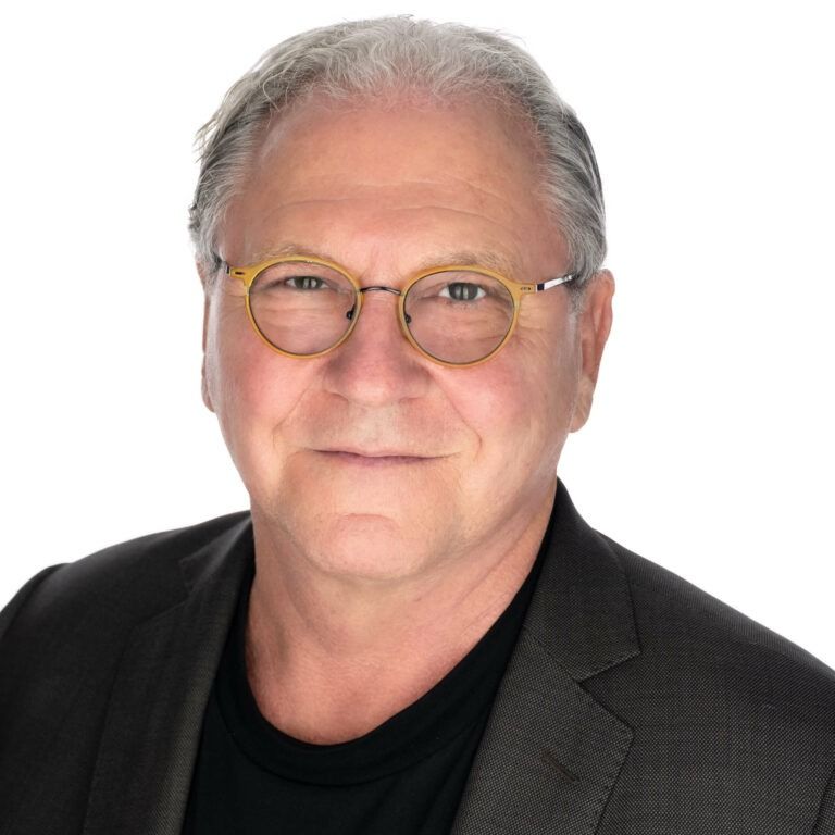 A person with short grey hair and round-framed glasses wearing a dark blazer over a black shirt against a white background.