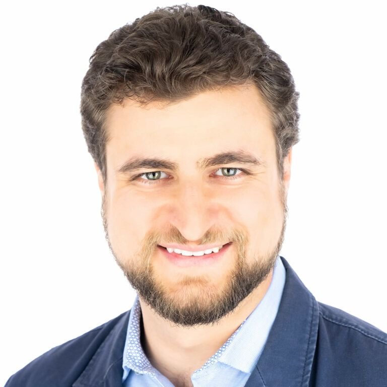 Headshot of a person with a short beard and brown hair, smiling and wearing a blue blazer over a patterned light shirt.