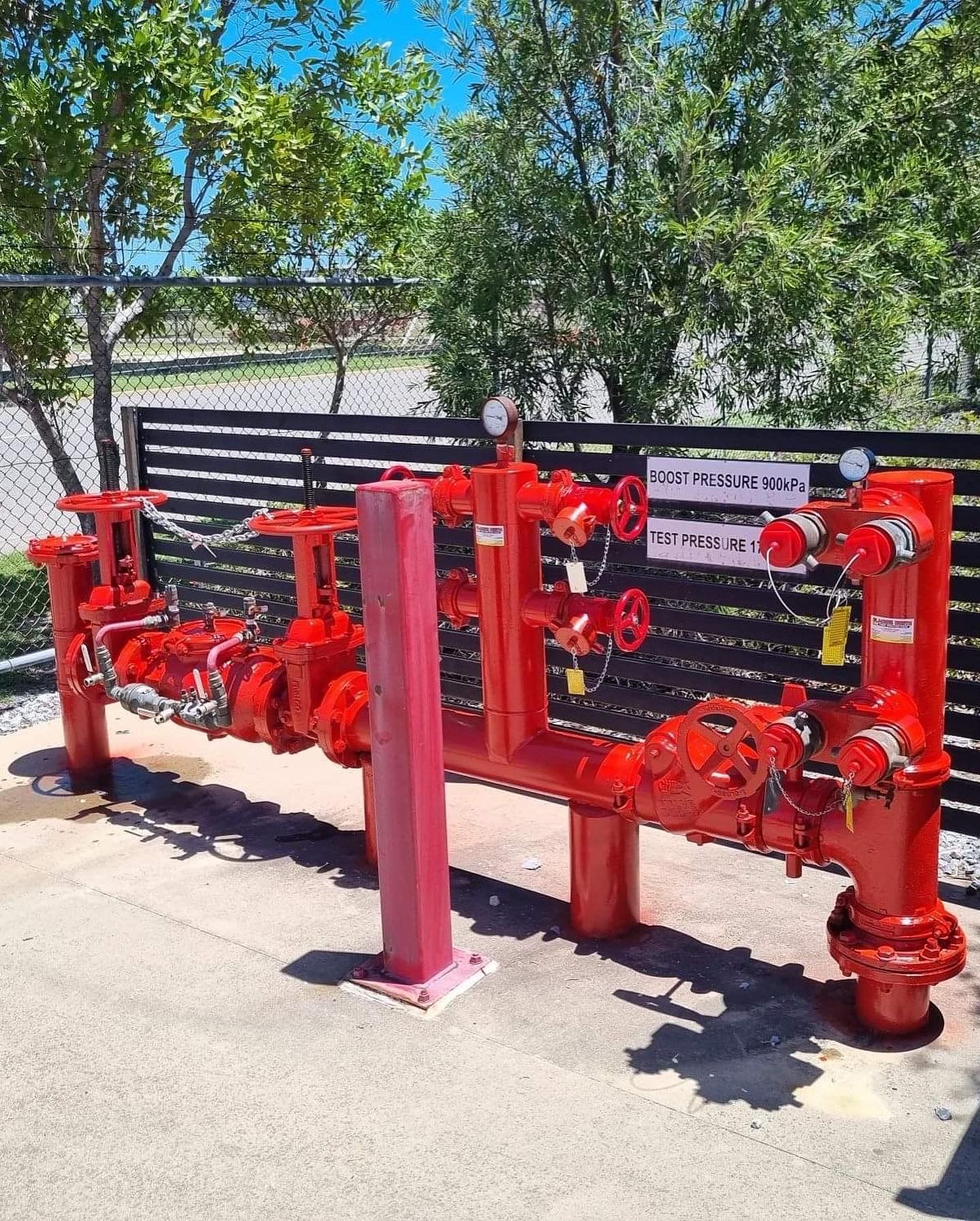 Red Fire Sprinkler Valves and Pipes Outdoors Beside a Fence and Trees — Flaming North Fire Protection Pty Ltd in Garbutt, QLD