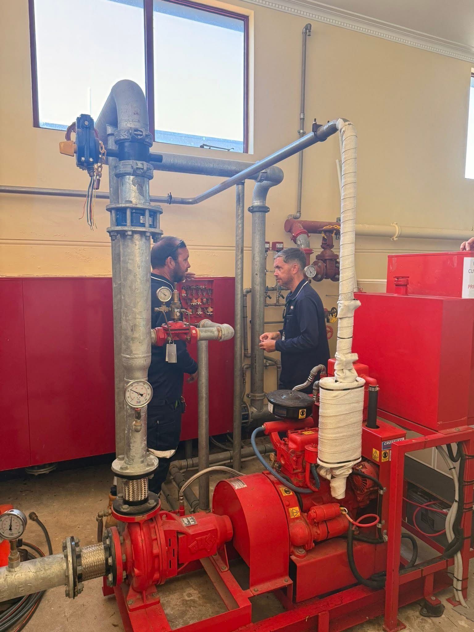 Two Workers Inspecting Red Industrial Pumps and White Pipes in a Boiler Room — Flaming North Fire Protection Pty Ltd in Garbutt, QLD