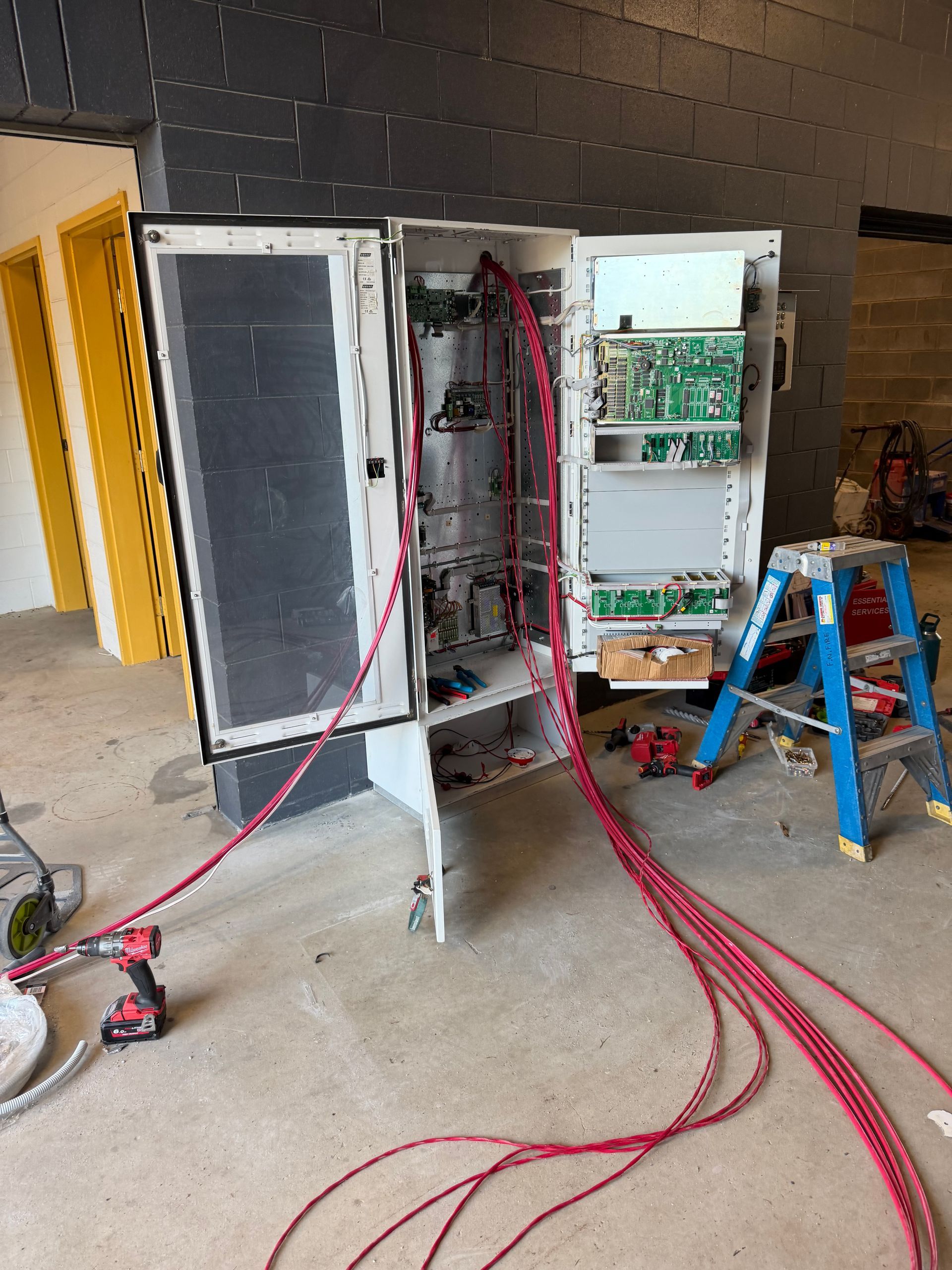 Electrical Cabinets With Exposed Wiring and Red Cables on a Workshop Floor, Beside a Blue Step Ladder — Flaming North Fire Protection Pty Ltd in Garbutt, QLD