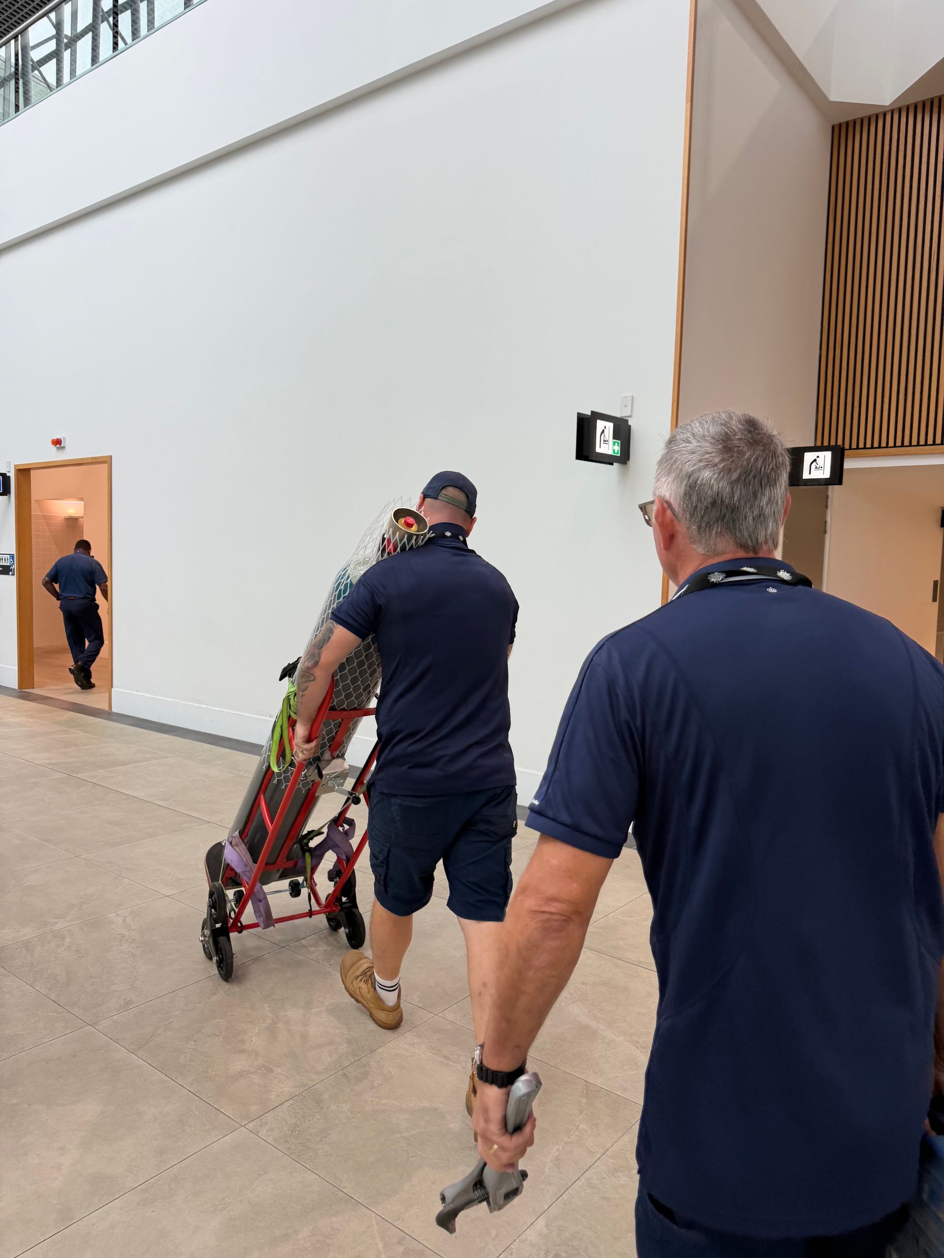 Two Men in Navy Shirts Carrying a Folded Stretcher Down a Hallway Toward an Open Door — Flaming North Fire Protection Pty Ltd in Garbutt, QLD