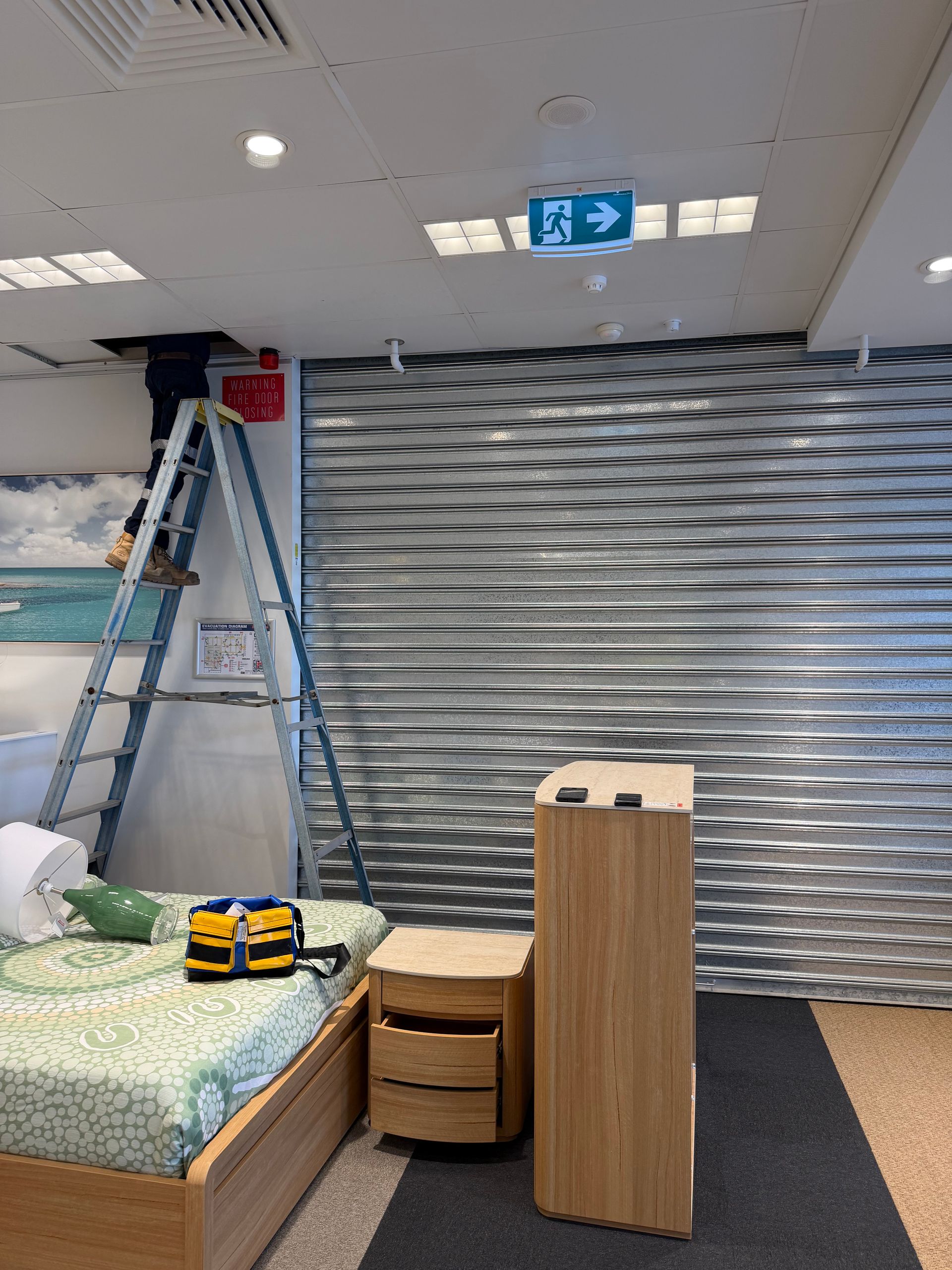 Small Room With a Ladder, Folded Metal Shutter, Bed, and Wooden Cabinets Under Ceiling Lights — Flaming North Fire Protection Pty Ltd in Charters Towers, QLD