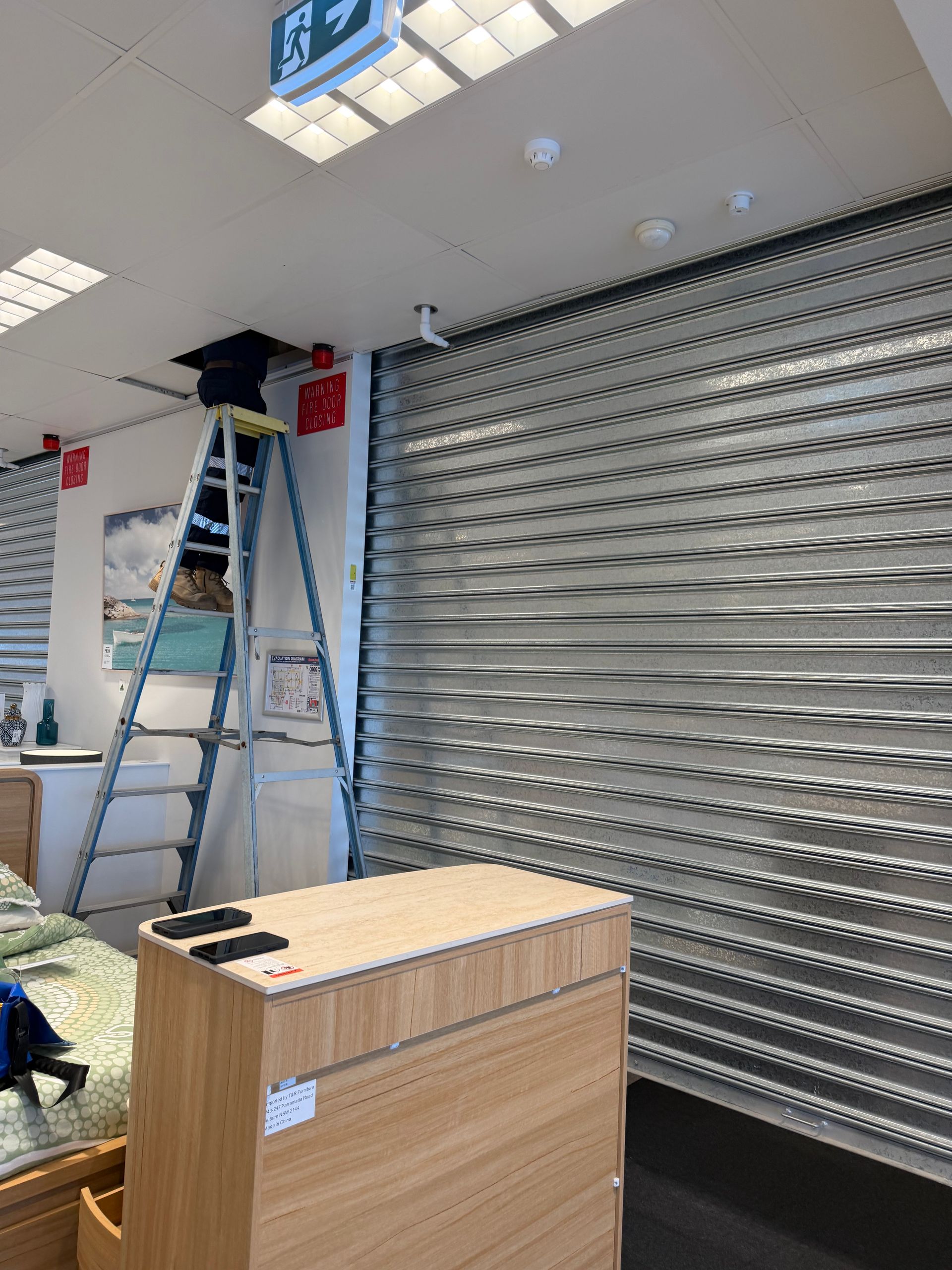 Indoor Storefront With a Metal Shutter, Ladder, and Wooden Cabinet Under Bright Ceiling Lights — Flaming North Fire Protection Pty Ltd in Garbutt, QLD