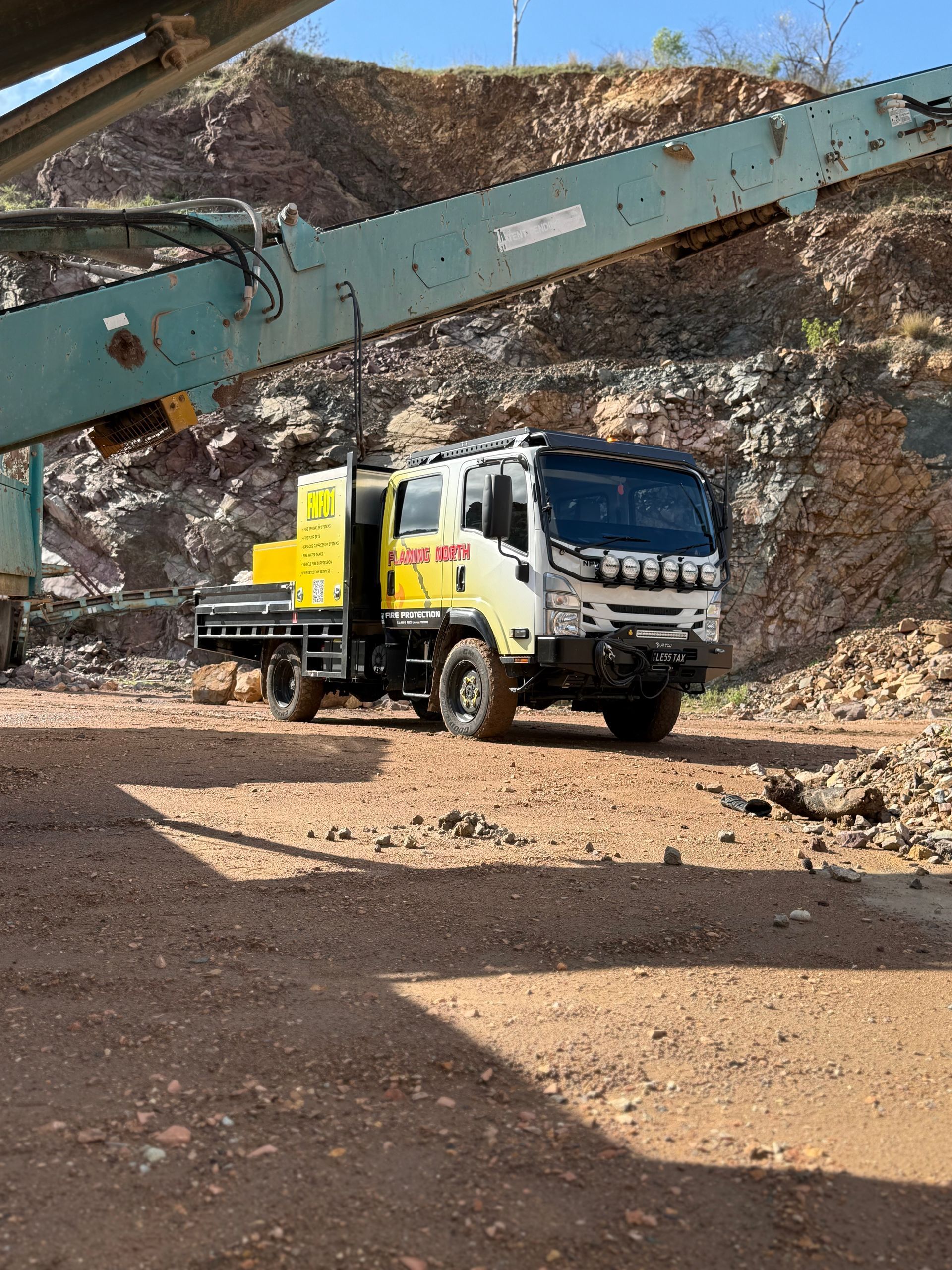 Yellow Utility Truck Parked in a Rocky Quarry Beneath a Large Conveyor Belt Structure — Flaming North Fire Protection Pty Ltd in Garbutt, QLD