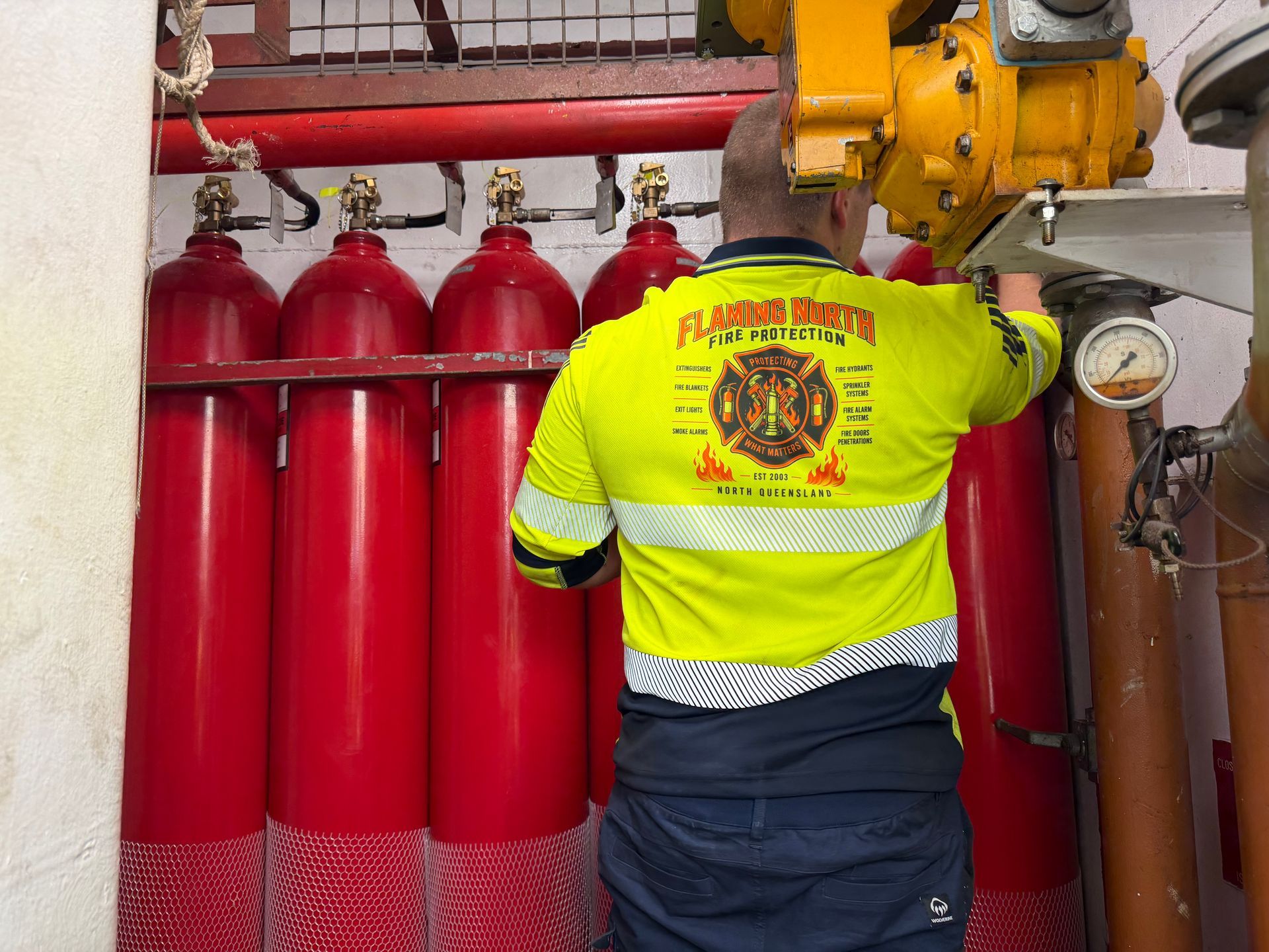 Worker In a High-visibility Jacket Inspecting Red Fire Suppression Cylinders and Pressure Gauges in an Industrial Room — Flaming North Fire Protection Pty Ltd in Mount Isa, QLD