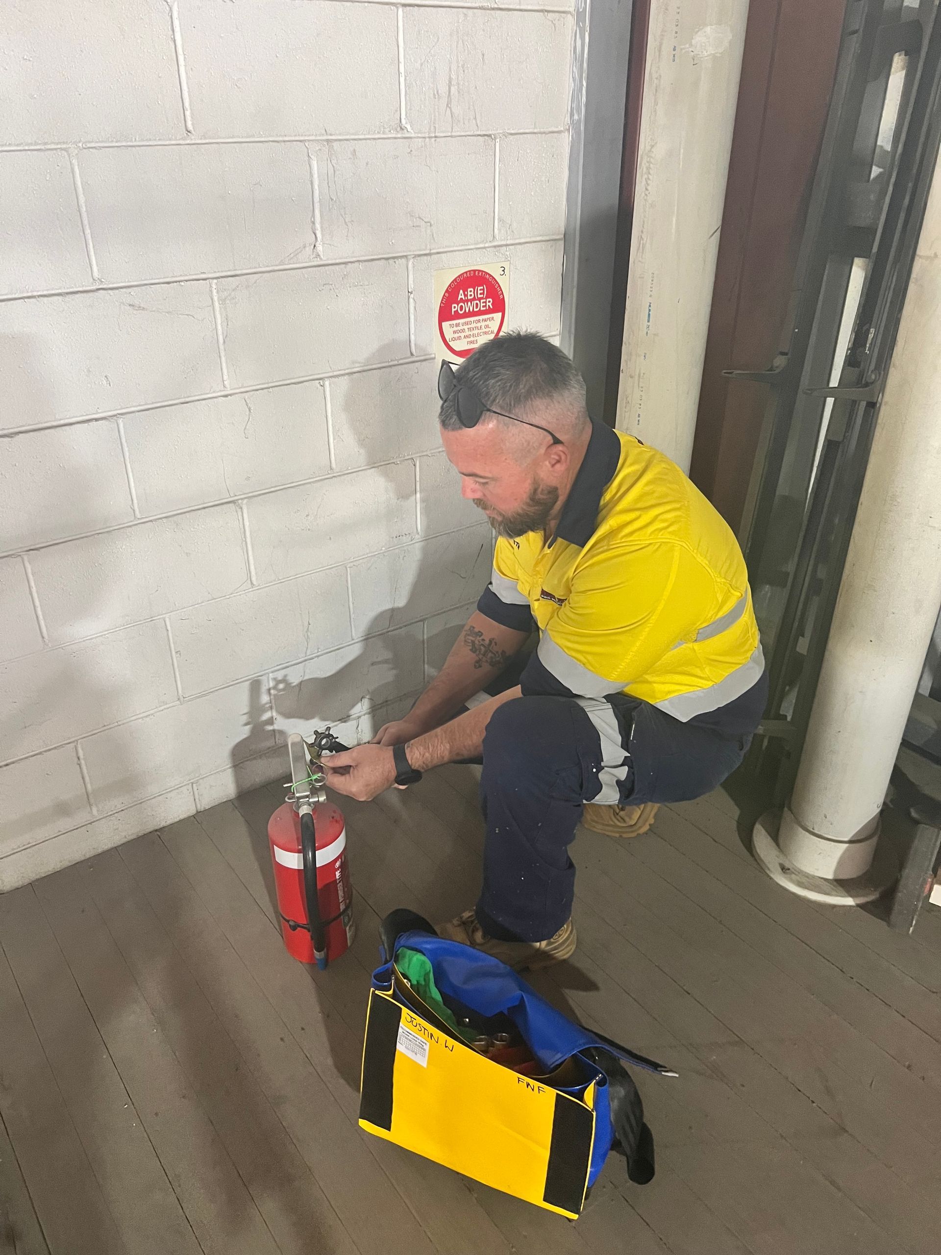 Worker In Yellow Shirt Crouches Beside a Red Fire Extinguisher in an Industrial Hallway — Flaming North Fire Protection Pty Ltd in Garbutt, QLD