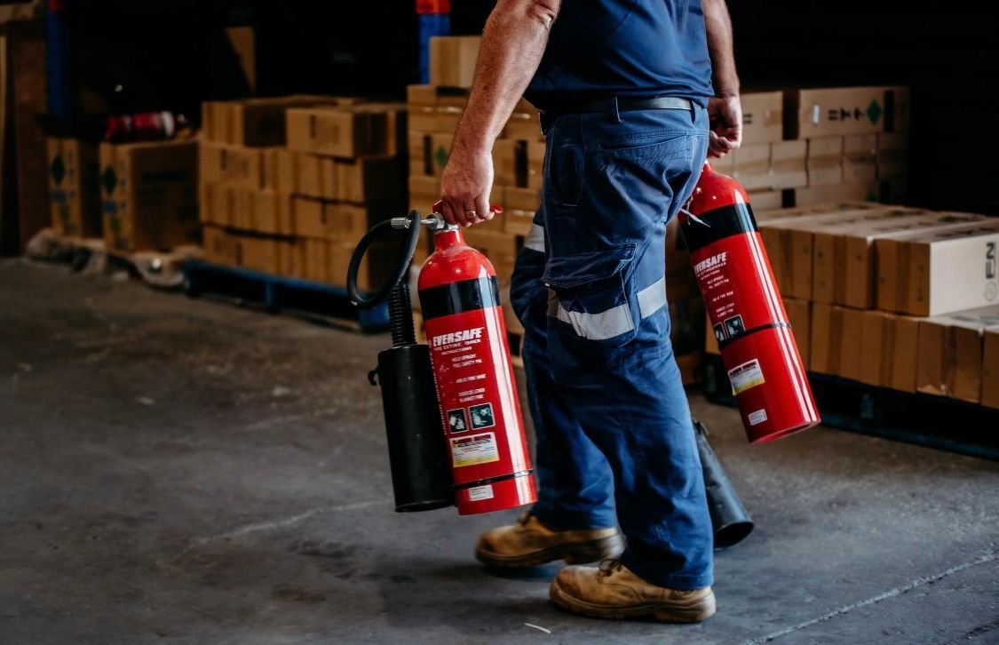 Worker Carrying Two Red Fire Extinguishers in a Warehouse Aisle — Flaming North Fire Protection Pty Ltd in Cairns, QLD