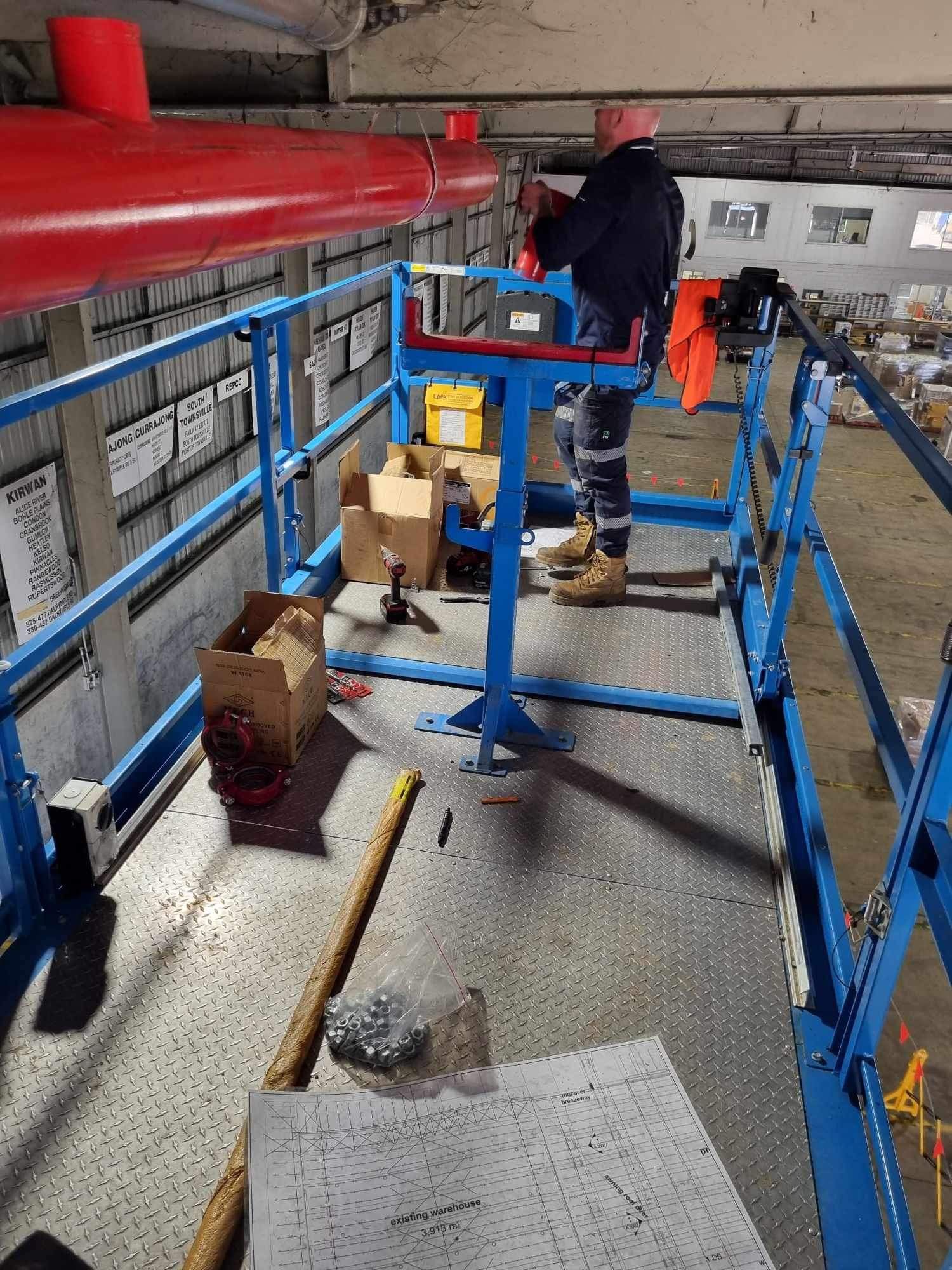 Worker On a Blue Industrial Platform Beside Red Pipes, Holding a Box in a Mechanical Room — Flaming North Fire Protection Pty Ltd in Garbutt, QLD