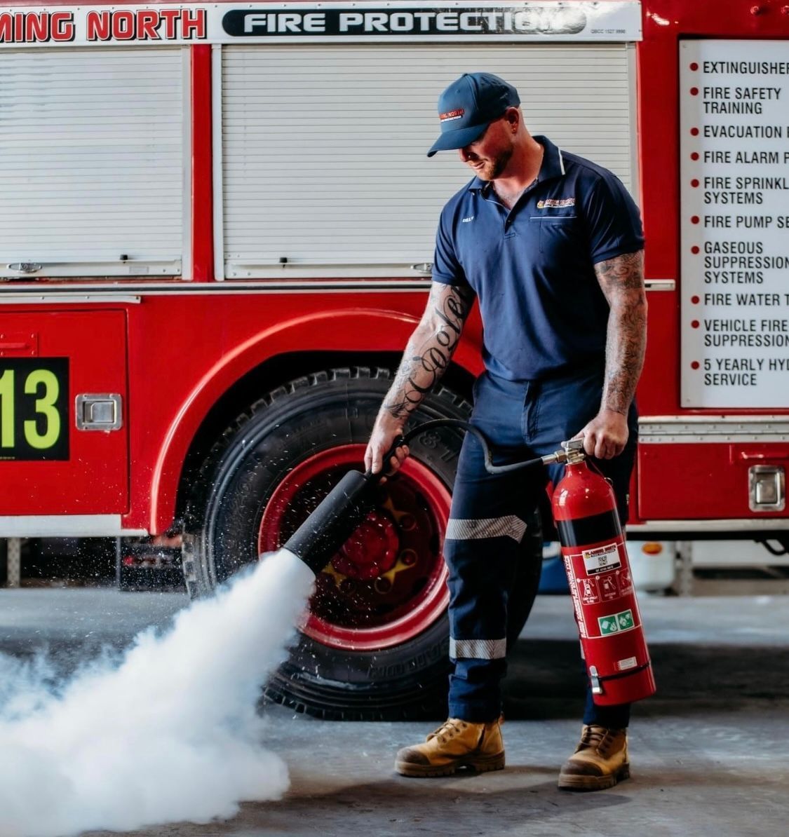 Firefighter In Navy Uniform Sprays Water From a Hose Beside a Red Fire Truck — Flaming North Fire Protection Pty Ltd in Garbutt, QLD